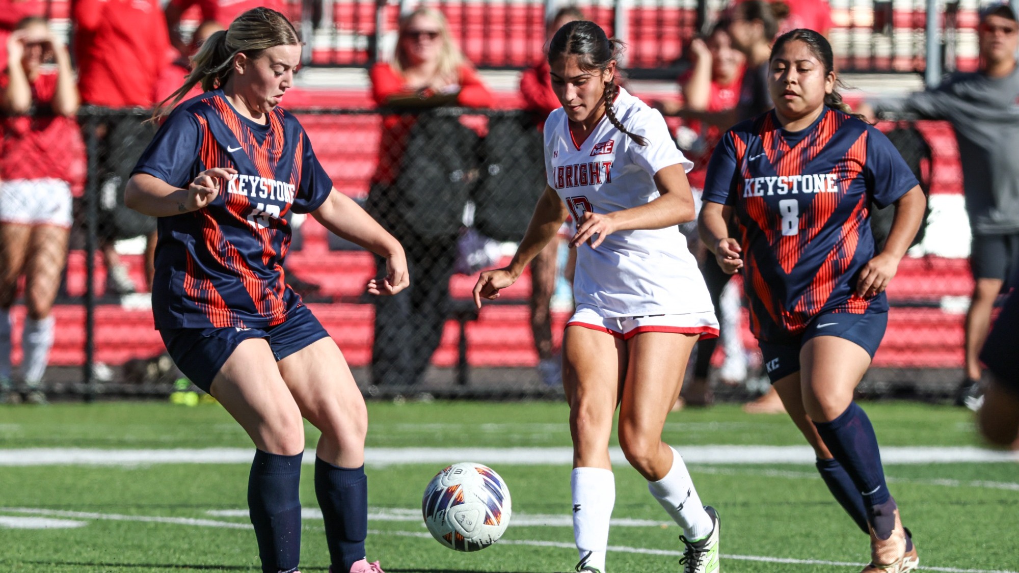 Two Keystone women’s soccer players in navy and orange uniforms challenge an Albright player in white for the ball, with spectators in red bleachers behind them.