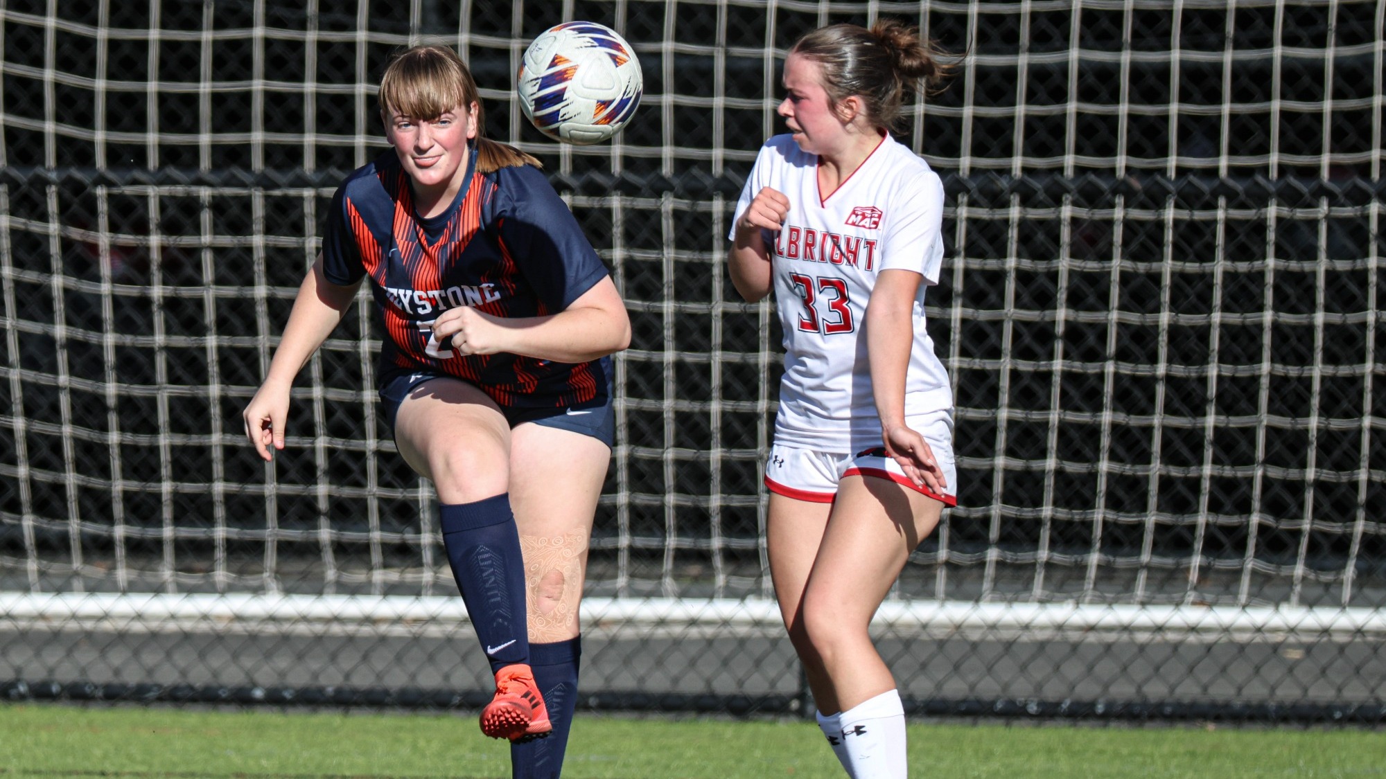 Keystone women’s soccer player in a navy and orange uniform raises her leg toward an airborne ball as an Albright player in white challenges beside her near the goal.