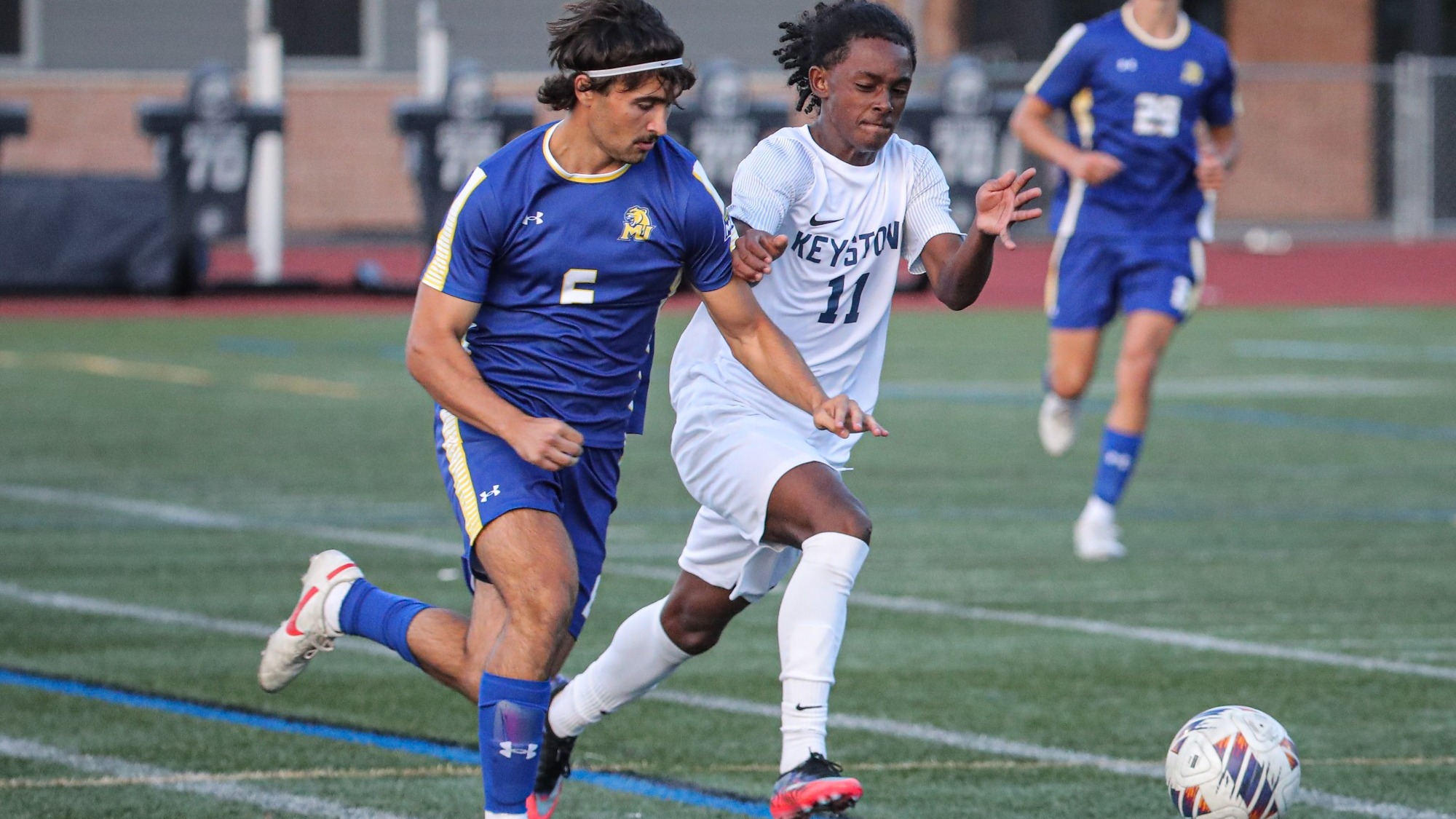Keystone men’s soccer player wearing a white No. 11 uniform runs alongside an opponent in a blue No. 5 uniform as both pursue the ball on the turf field.