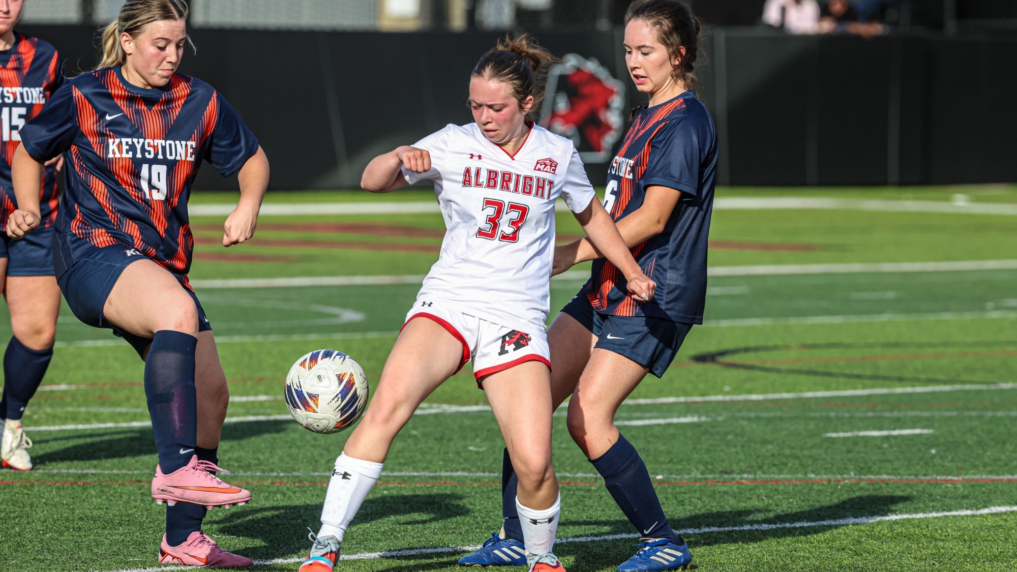 Keystone women’s soccer players in navy and orange uniforms challenge Albright’s No. 33 for the ball on a turf field, with Keystone’s No. 19 lifting her foot toward the ball.