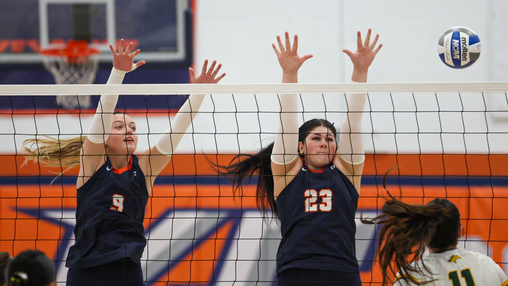 Two Keystone College volleyball players jump at the net to block an opposing attacker during a match, with a volleyball visible in the upper right corner.