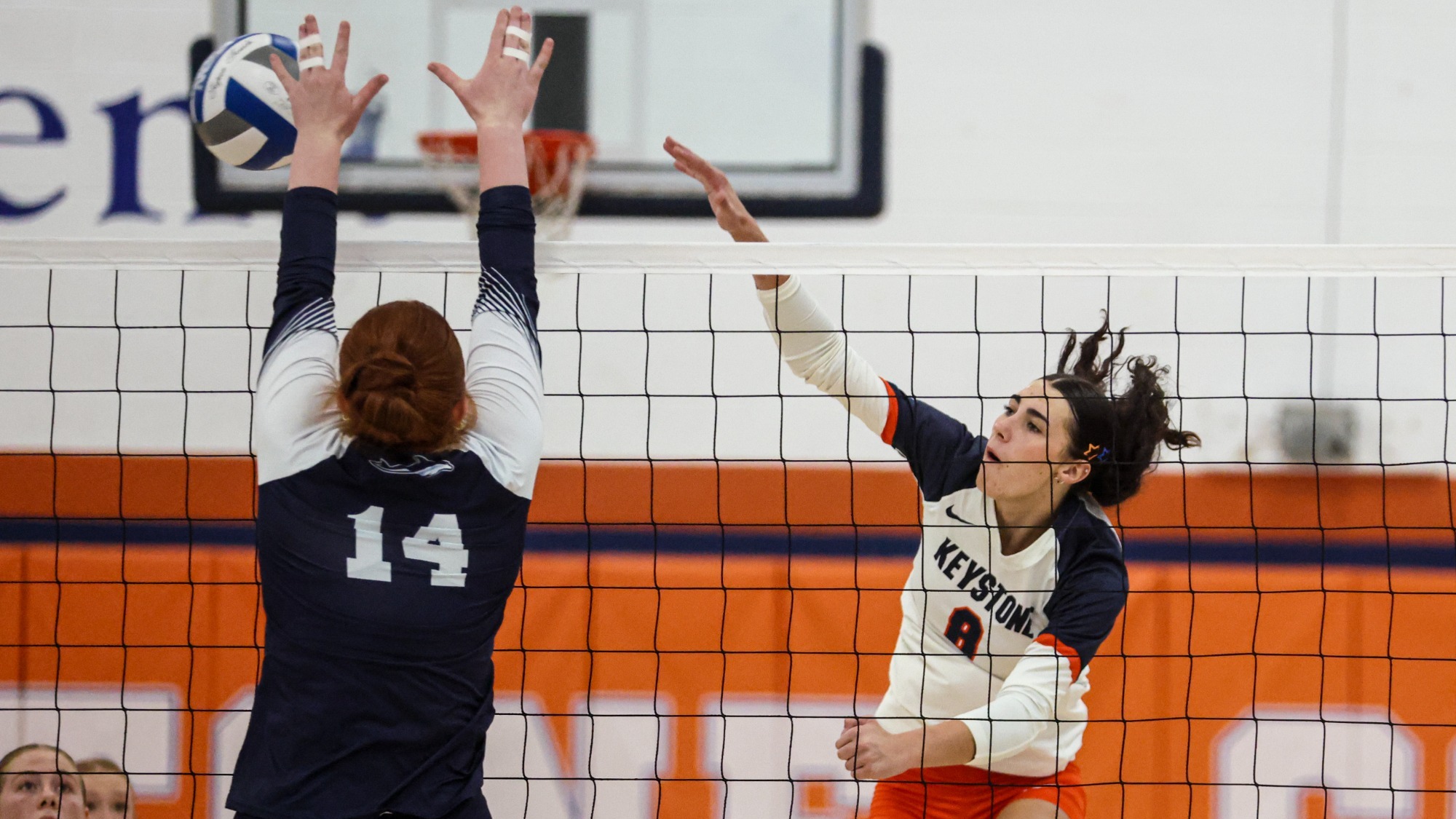 A volleyball player in a white and orange uniform spikes the ball over the net as an opposing player in navy attempts to block it. Teammates watch from behind the net.