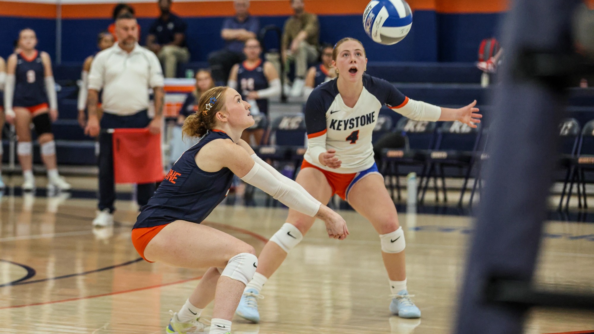 Two Keystone College volleyball players prepare to receive the ball during a match. One player is crouched low with her arms extended, while a teammate stands behind her watching the ball in the air. The Keystone team is visible in the background on the court.