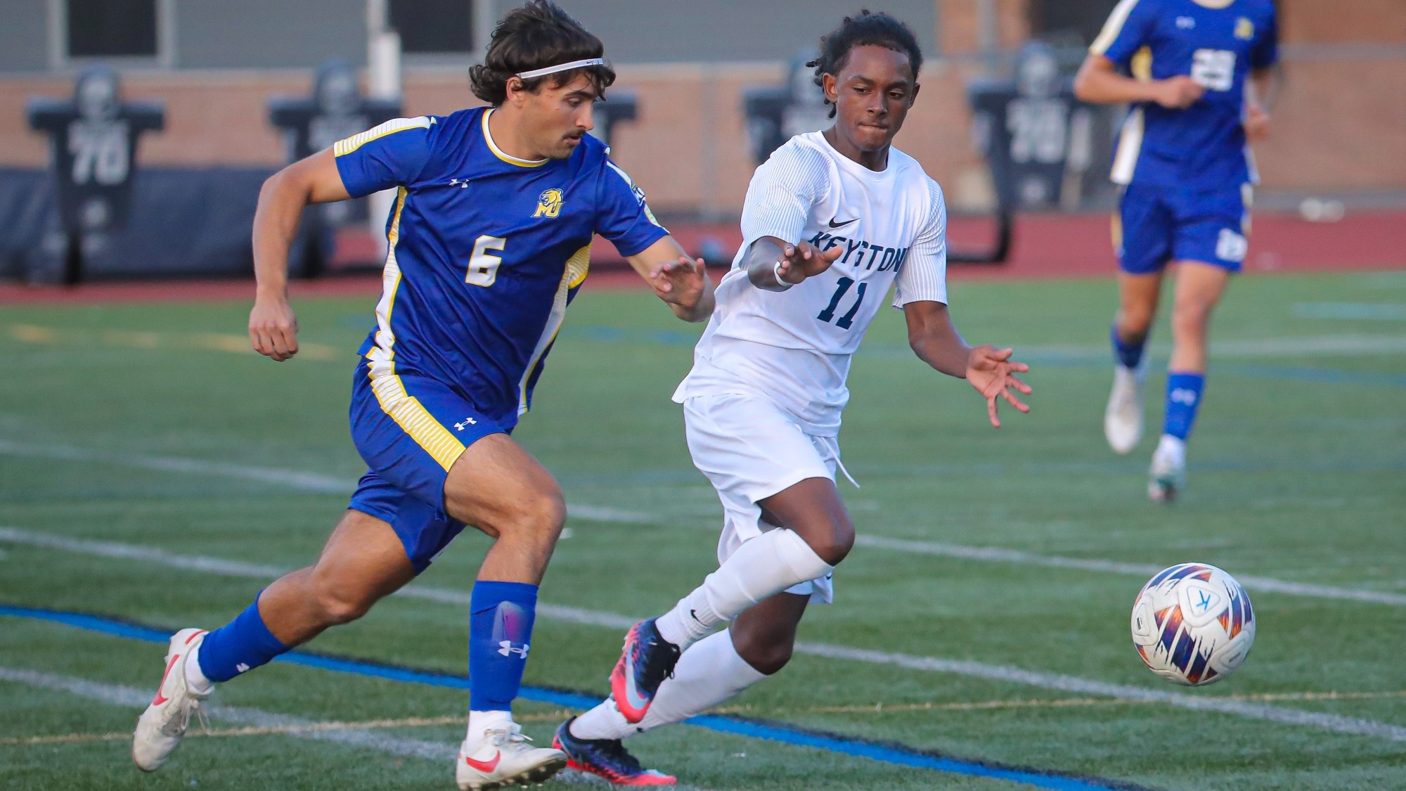 Keystone College men’s soccer player Dary Hodge-Davila, wearing No. 11, races alongside an Elmira player while pursuing the ball during a road match. Another Keystone player is visible in the background.