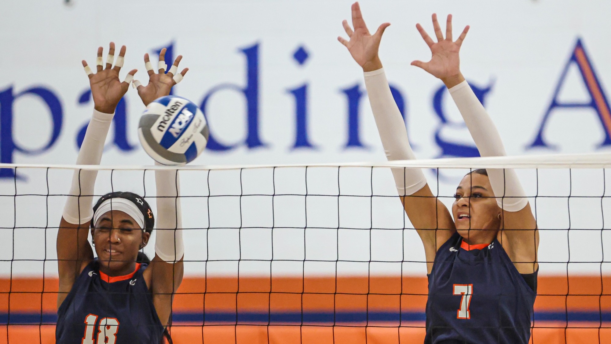 Keystone volleyball players #18 and #7 battle at the net, jumping with arms extended to block an opposing attack as the volleyball hangs just above the net. Both players wear Keystone uniforms, with a gymnasium backdrop featuring blue and orange accents.