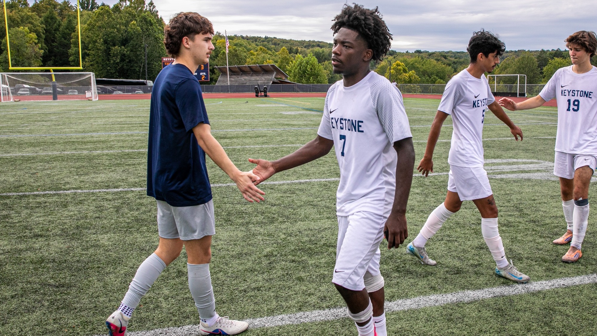 Keystone College men’s soccer players shake hands on the field following a game, with player No. 7 in white at center and teammates in the background.
