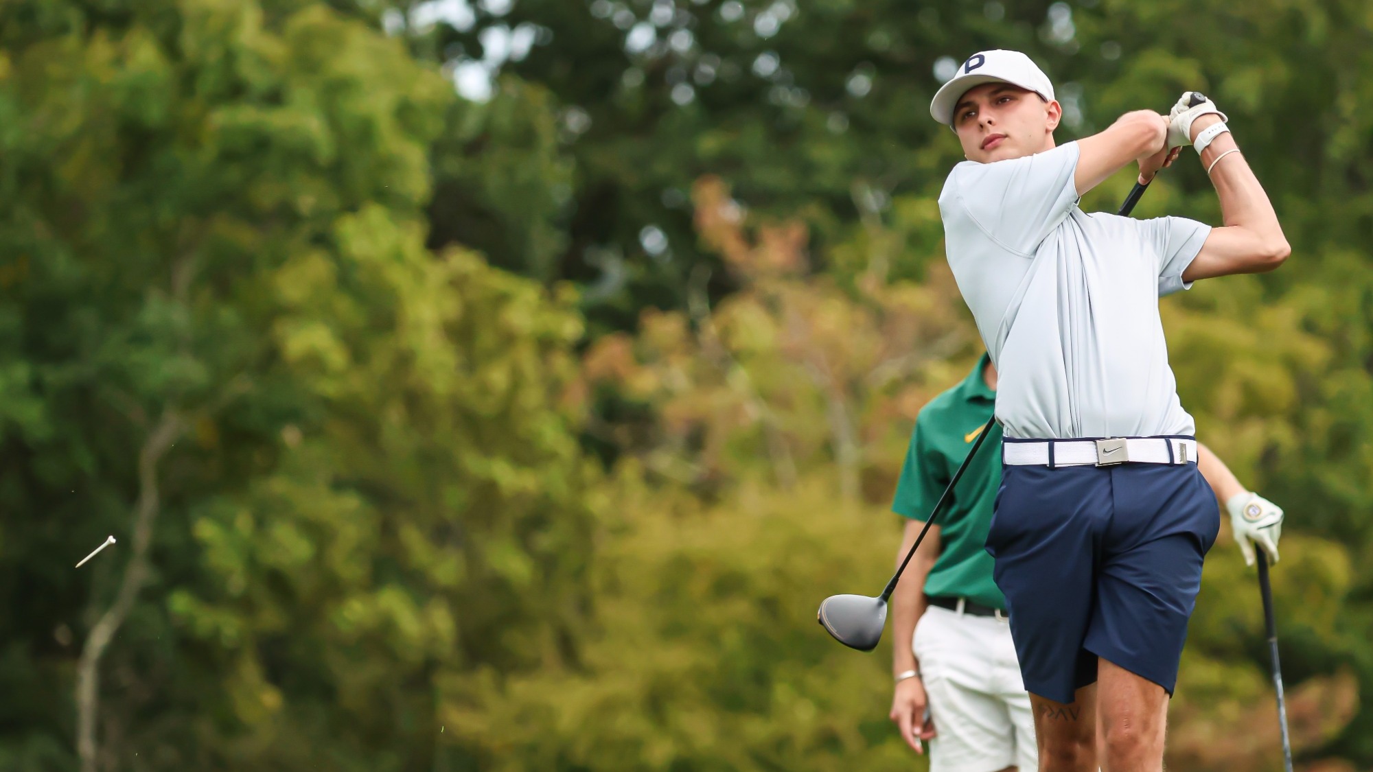 Keystone College men’s golfer follows through on a tee shot during a round, with a wooded course in the background and another golfer standing behind him.