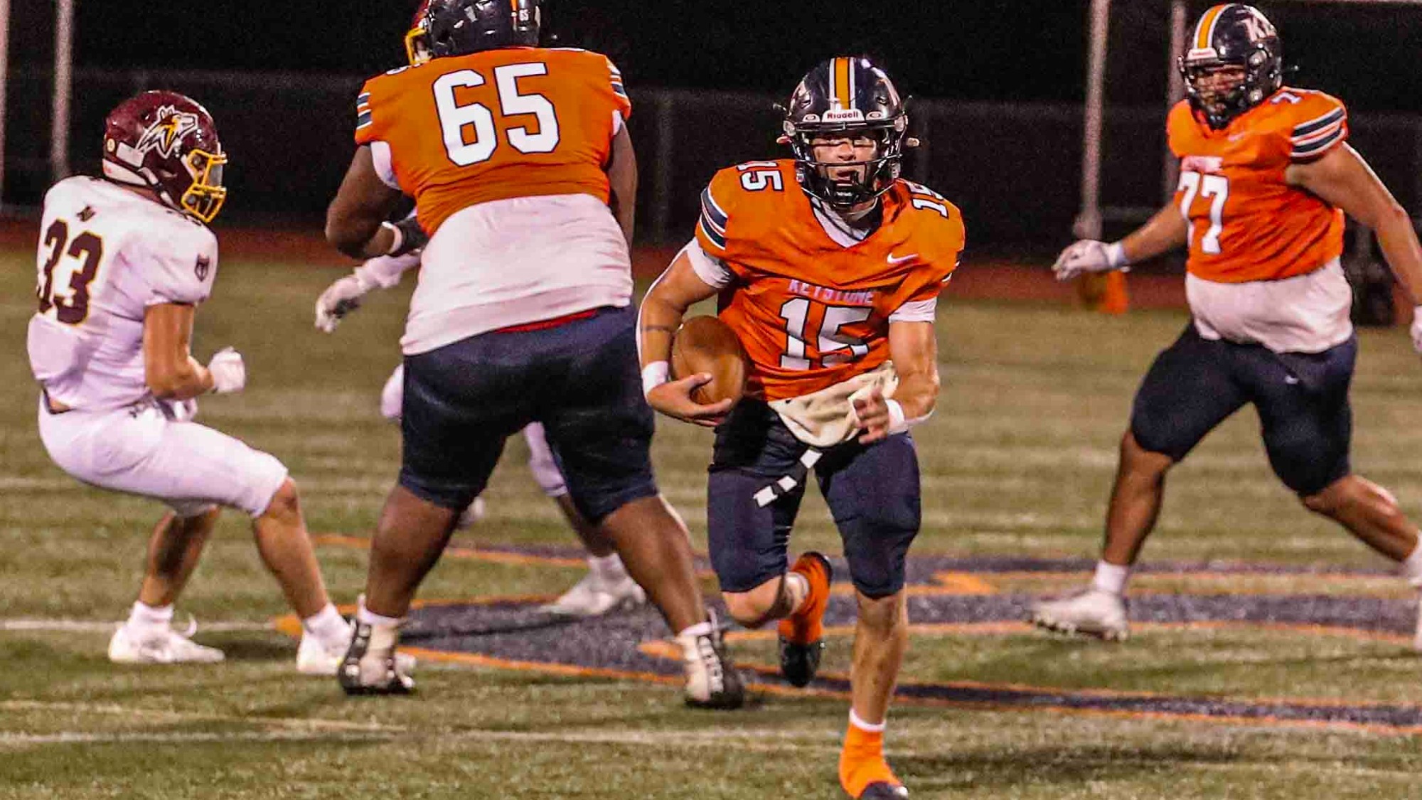 Keystone College football quarterback No. 15 carries the ball downfield as teammates No. 65 and No. 75 block defenders during a night game.