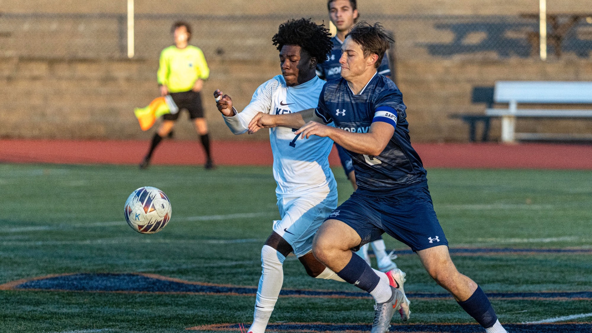 Keystone men’s soccer player in a light blue and white uniform battles for possession against an opposing player in a navy uniform, with the ball just ahead of them on the field.