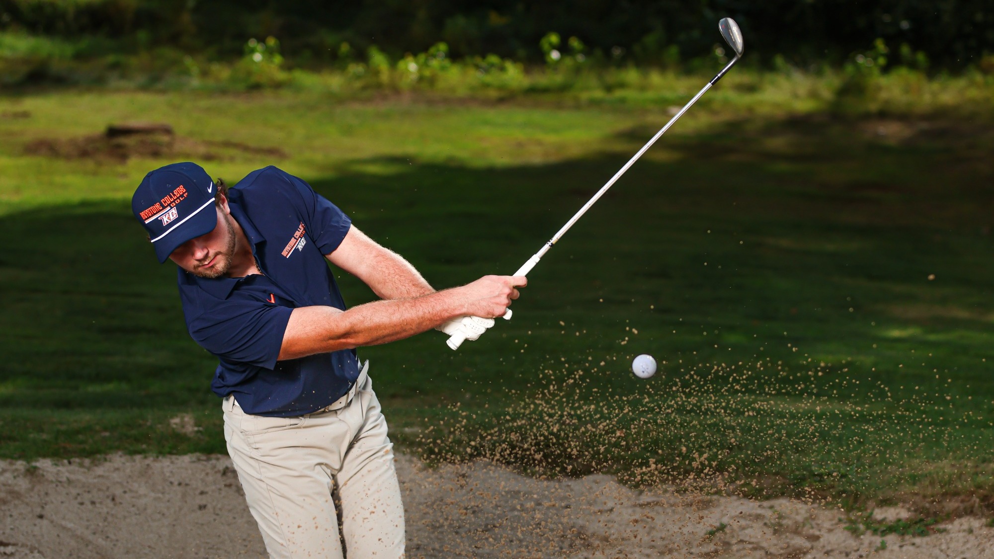 A Keystone men's golfer in a navy uniform and cap swings from a sand bunker, sending sand into the air as the golf ball travels forward. The golfer is captured mid-swing with a wooded, grassy course in the background.