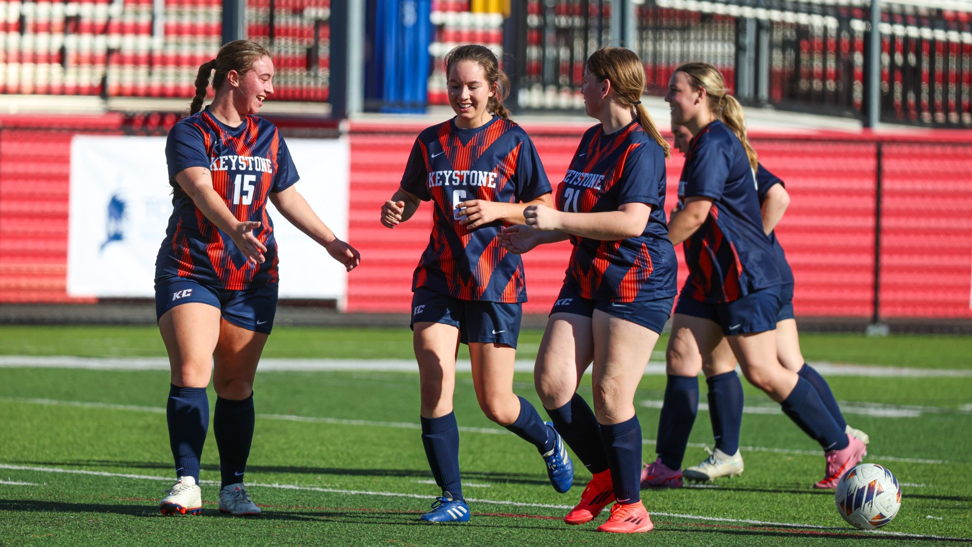Four Keystone women’s soccer players in navy and red uniforms run together on the field during a game, with a soccer ball in the foreground and red-and-white stadium seating in the background.
