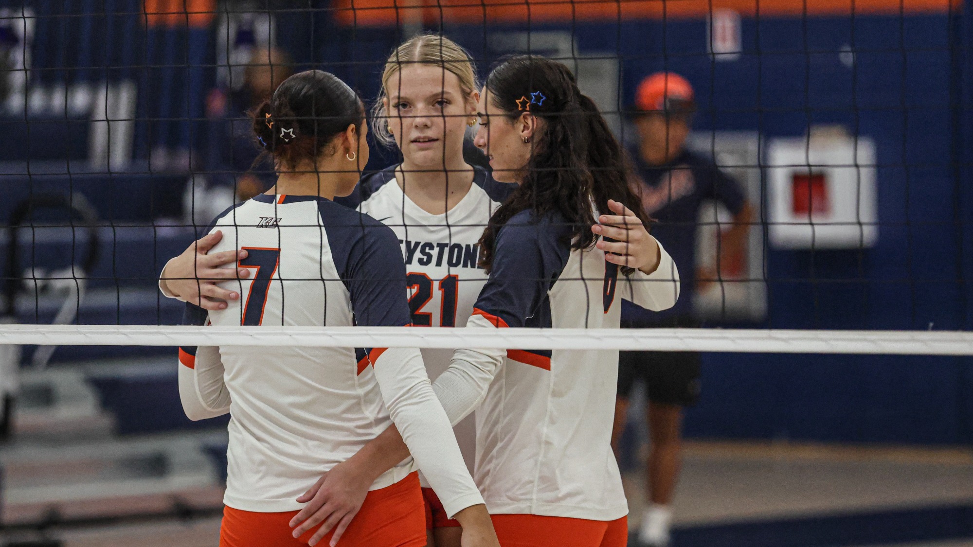 Three Keystone College volleyball players in white and orange uniforms gather together at the net during a match, with one player wearing No. 21 centered between two teammates as they regroup. The volleyball net runs across the foreground, with the court and spectators visible in the background.