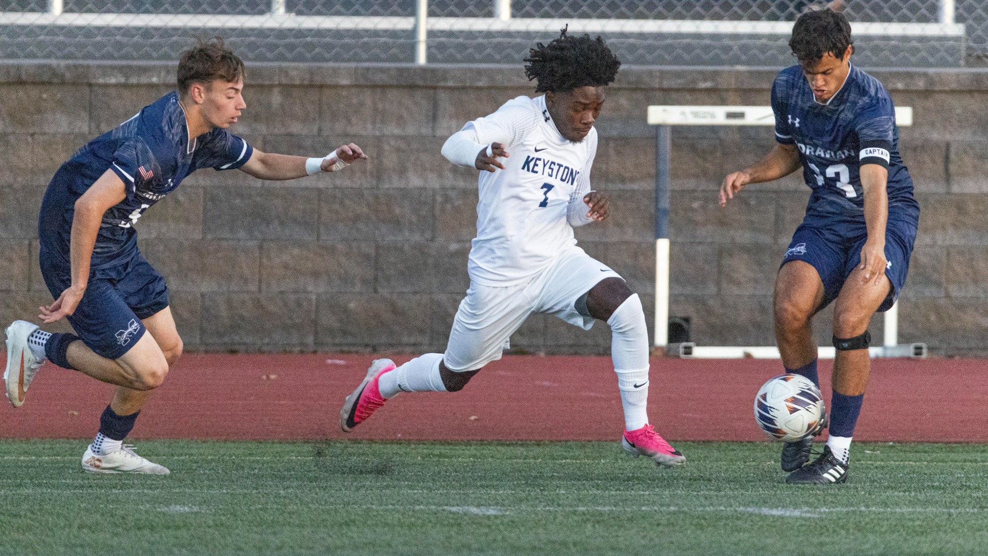 A Keystone soccer player in a white No. 7 jersey runs toward the ball as two opponents in navy defend.