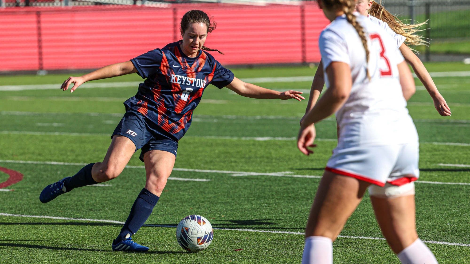A Keystone soccer player wearing No. 6 prepares to kick the ball as two players in white defend on the field.