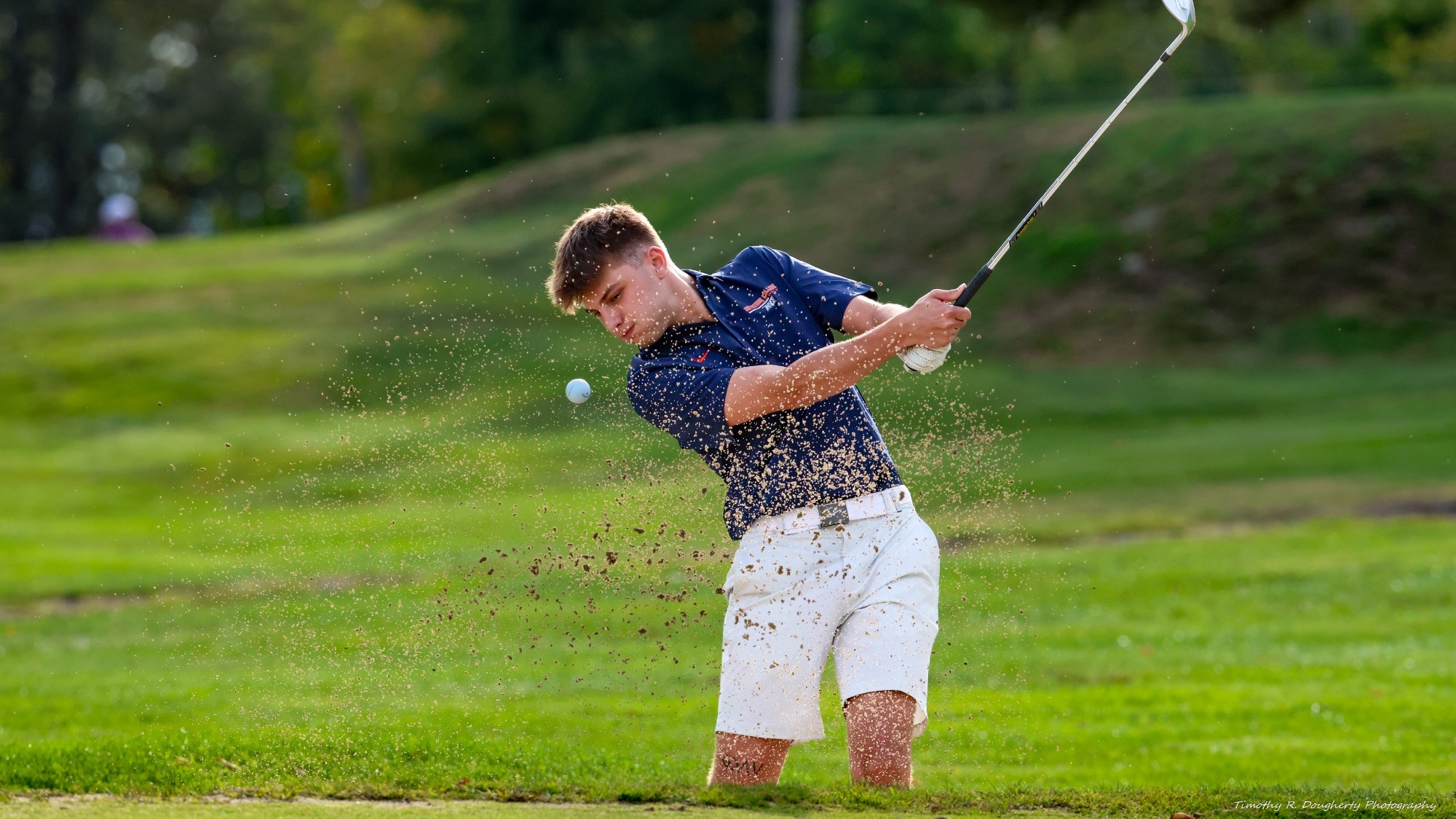 A golfer in a navy polo and white shorts hits a shot from a sand bunker, sending sand into the air as the ball flies toward the green.