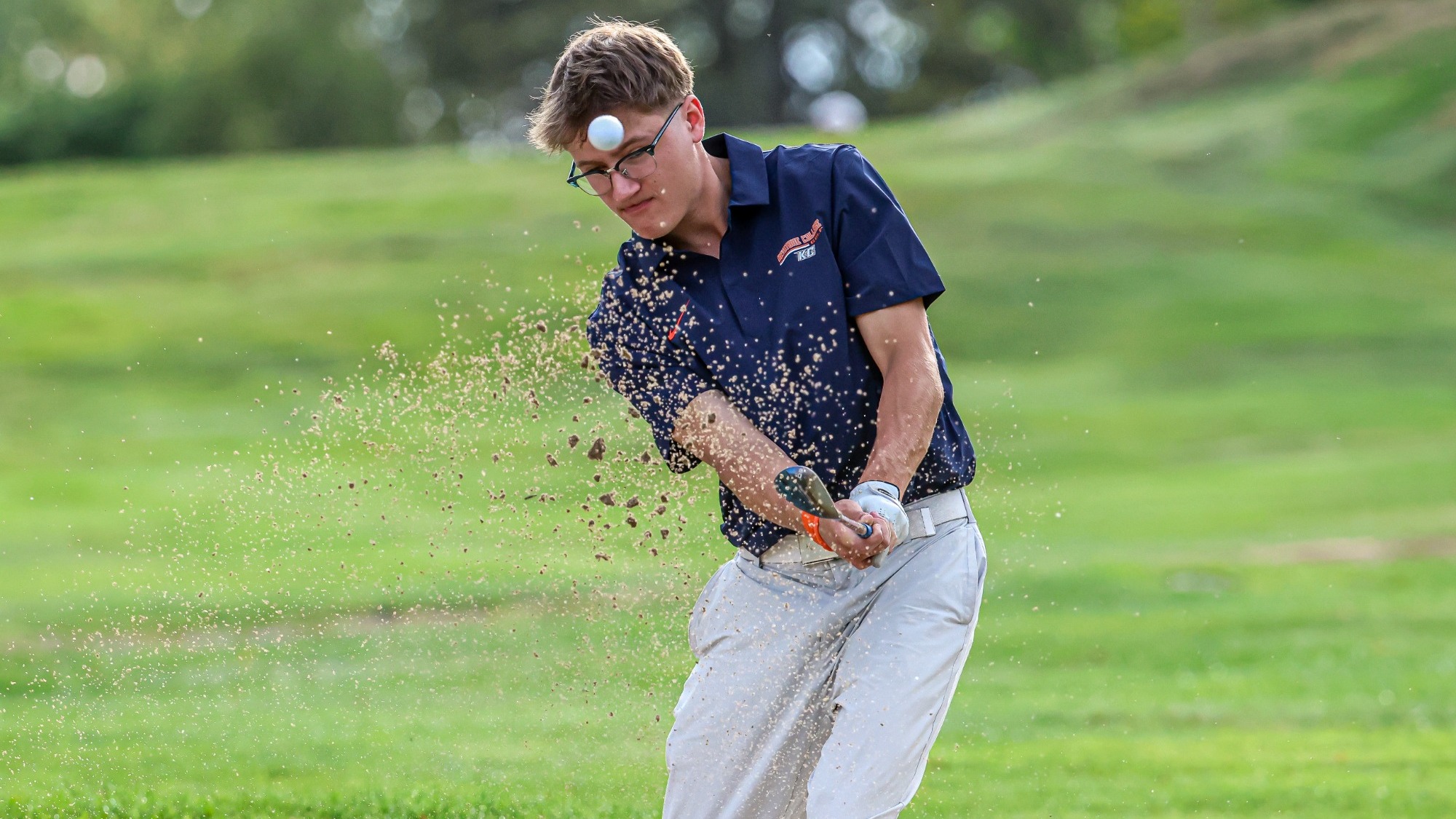A Keystone golfer in a navy polo and gray pants hits a shot from a sandy area. Sand sprays into the air as the ball rises in front of him.