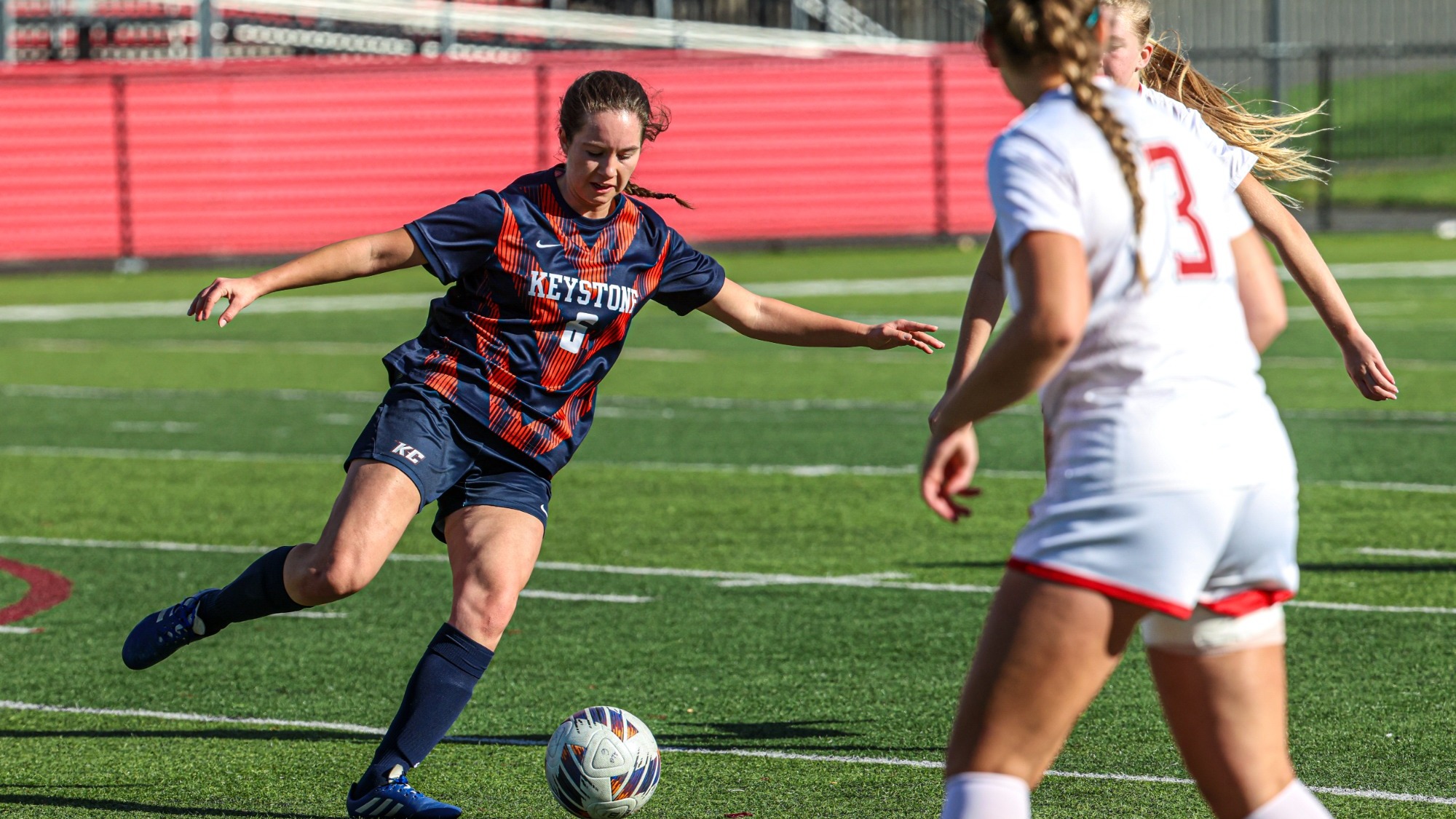 Keystone women’s soccer player wearing number 6 in a navy and orange uniform prepares to kick the ball as two opposing players in white approach on the turf field.