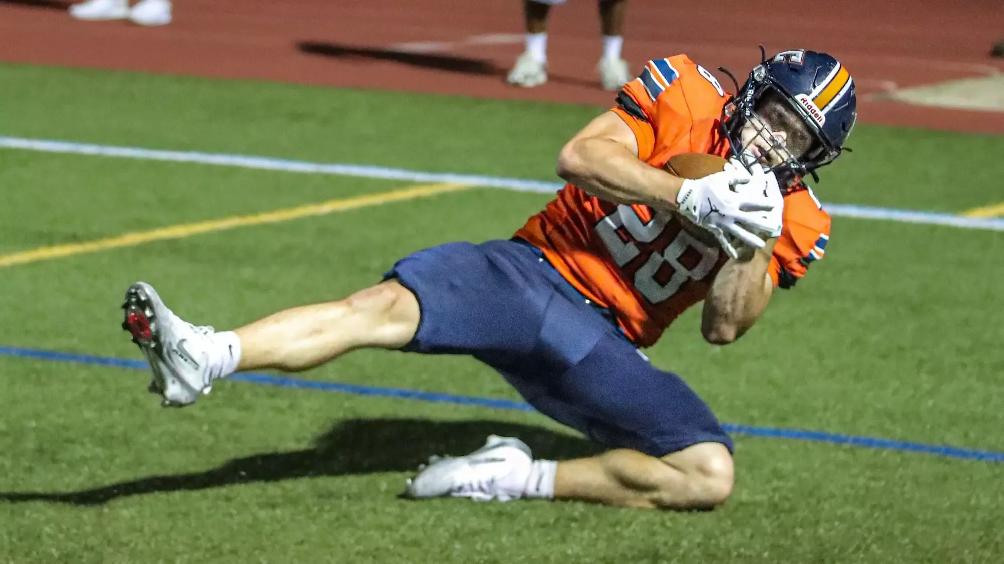 Keystone College football player in an orange uniform and helmet dives forward while securing the football during a game on the turf field.