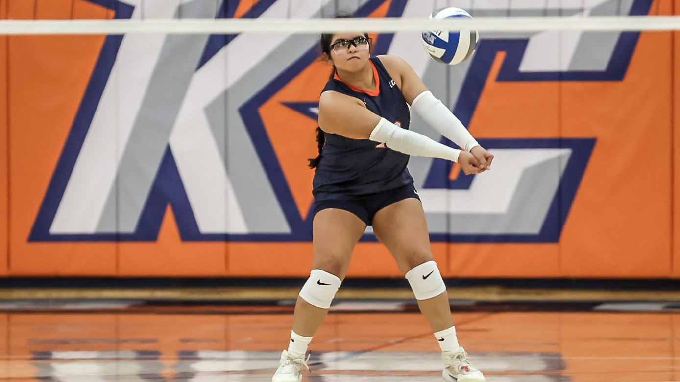 Keystone volleyball player preparing to receive the ball during a match, with the athlete wearing a navy and orange uniform, white arm sleeves and knee pads, positioned on the court in front of an orange-and-navy Keystone backdrop.