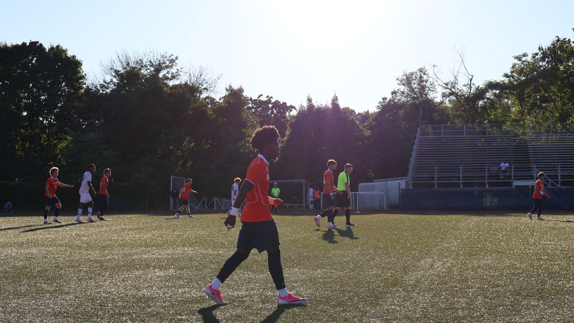 Keystone College men's soccer players compete on the field during a game, with a player in red in the foreground and teammates and opponents in the background.