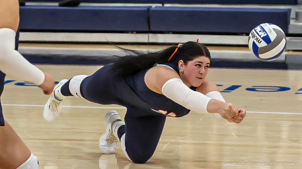 A volleyball player in a navy uniform dives forward across the court with her arms extended to bump the ball, while another player is partially visible nearby. A volleyball floats just ahead of her.