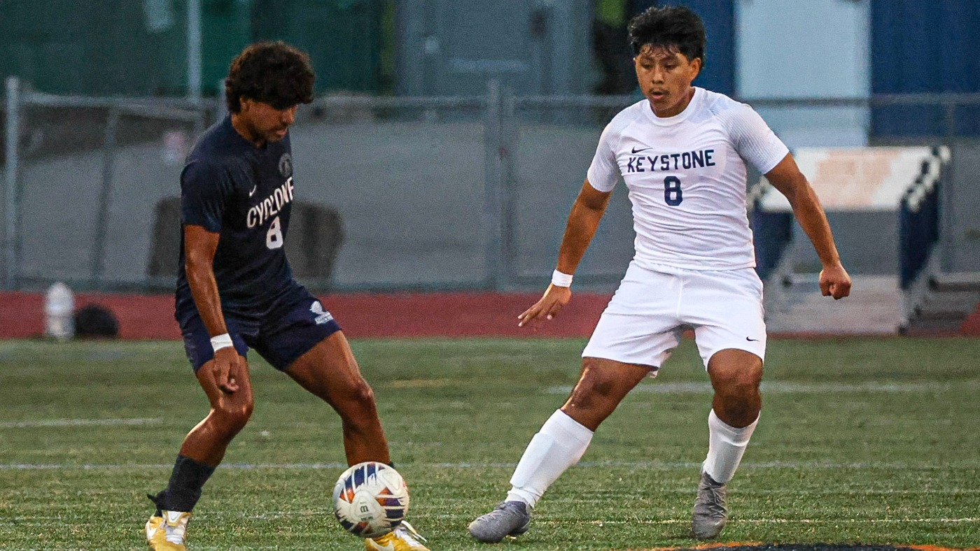 Keystone men's soccer player Joel Calle, wearing a white No. 8 uniform, battles for possession of the ball against a Centenary player in a navy No. 8 uniform during a match.