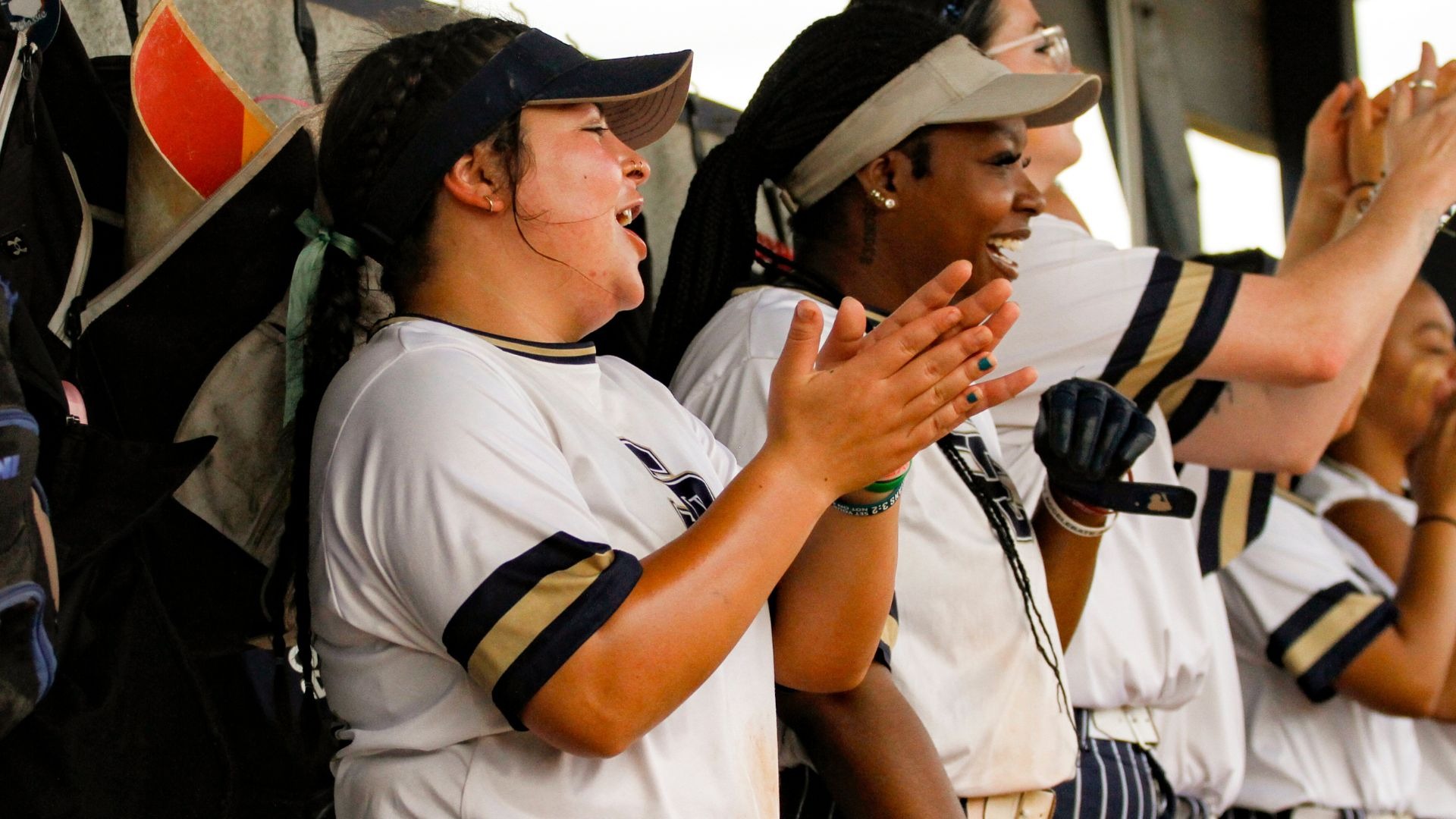Softball players clap while standing inside the dugout.