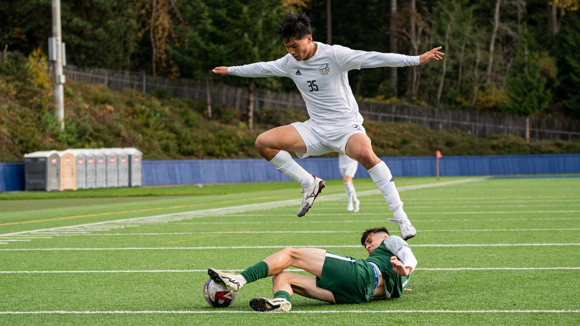Henri Godbout leaping over a UFV Cascades player on the ground, Godbout looking down at the ball at the UFV player's feet, arms and legs spread wide in a ninja-like position as he prepares to land