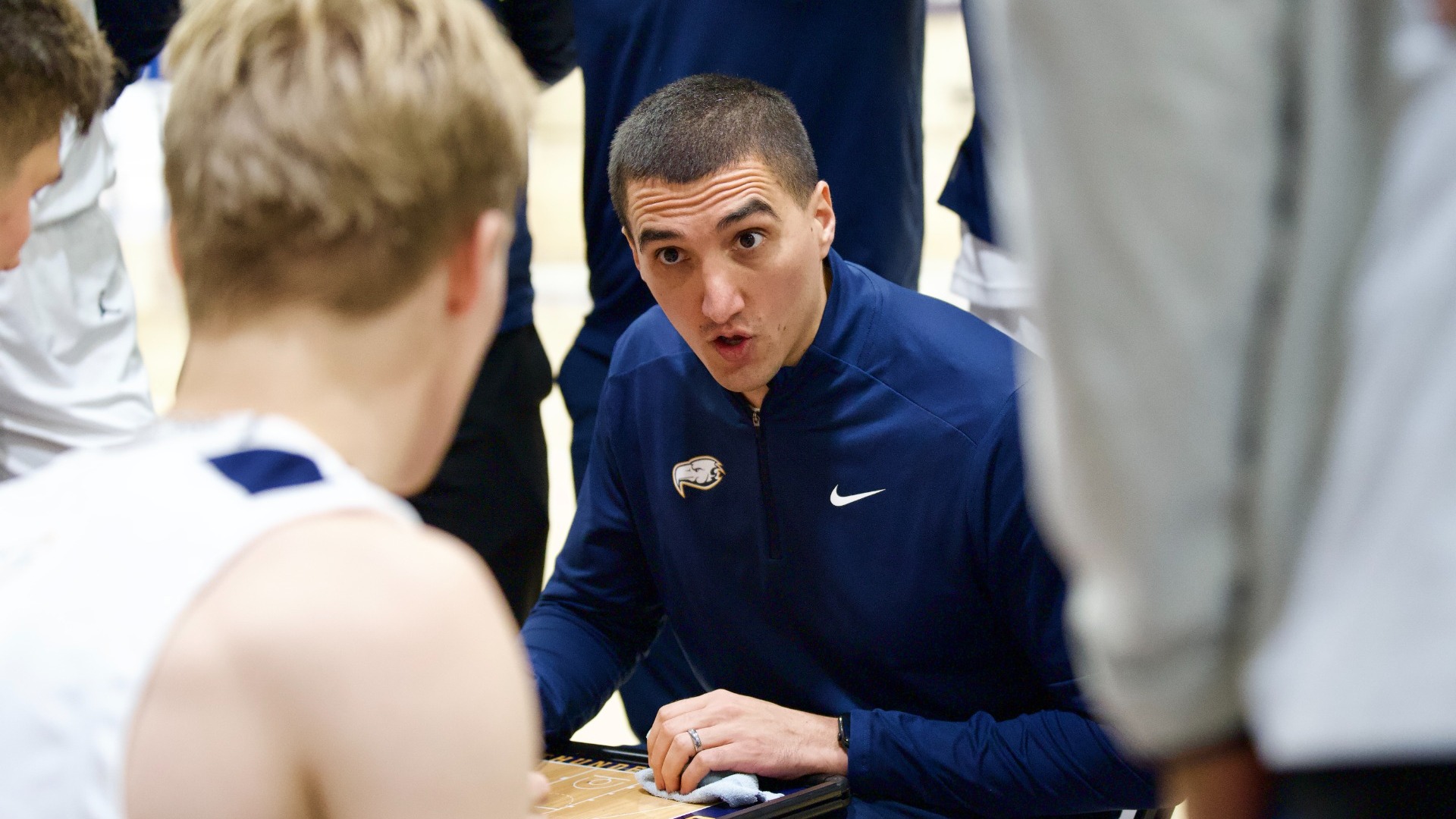 UBC men's basketball head coach Phil Jalalpoor seen in focus between two of his players, both out of focus in the foreground, explaining something intensely during a timeout