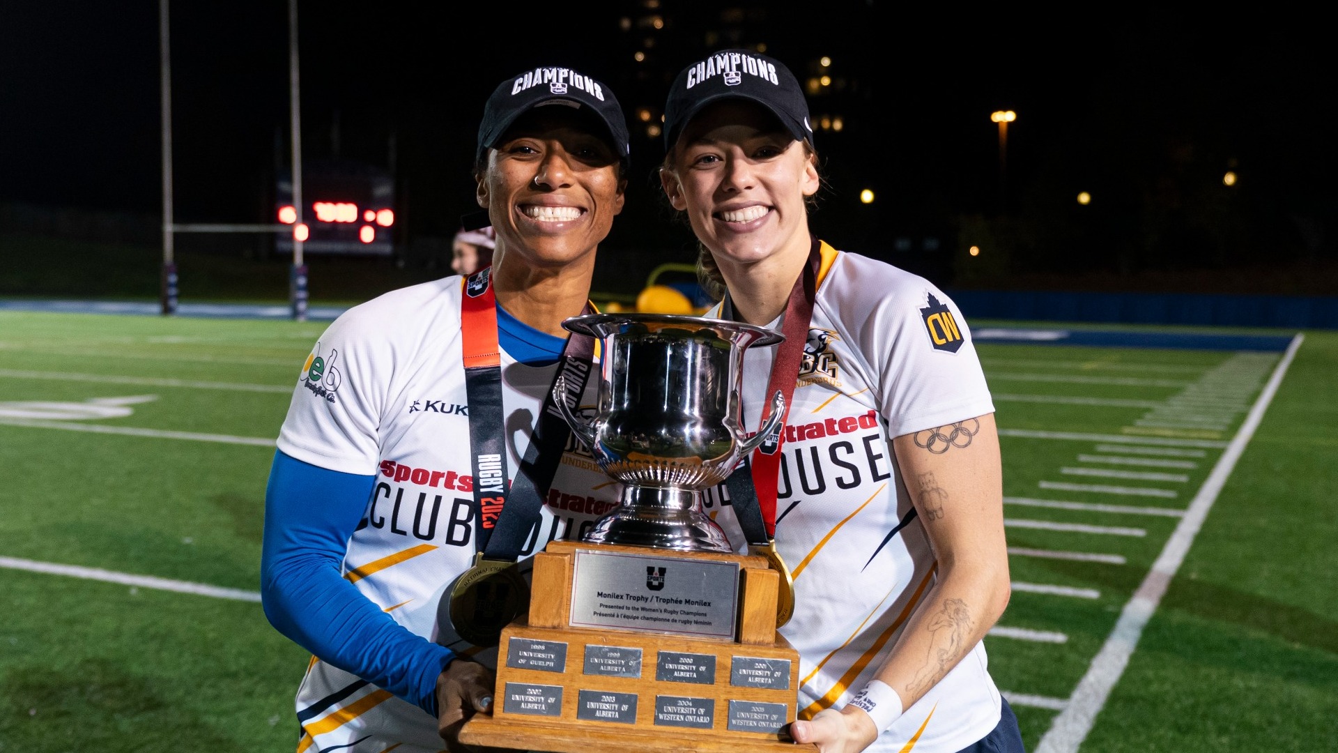 Piper Logan and Charity Williams smiling and posing with the U SPORTS Championship trophy after winning the 2025 Women's Rugby Championship
