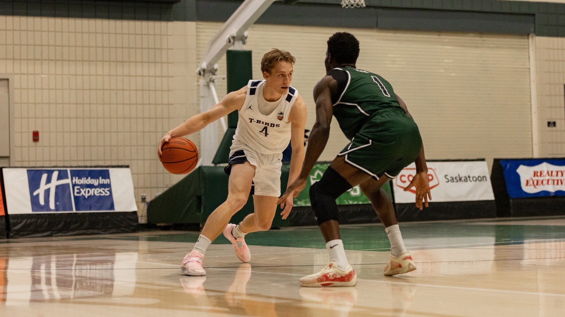 UBC's Holt Tomie dribbling the ball up the court, on his side of half, in the midst of a crossover with a Saskatchewan Huskies player squared up and guarding him