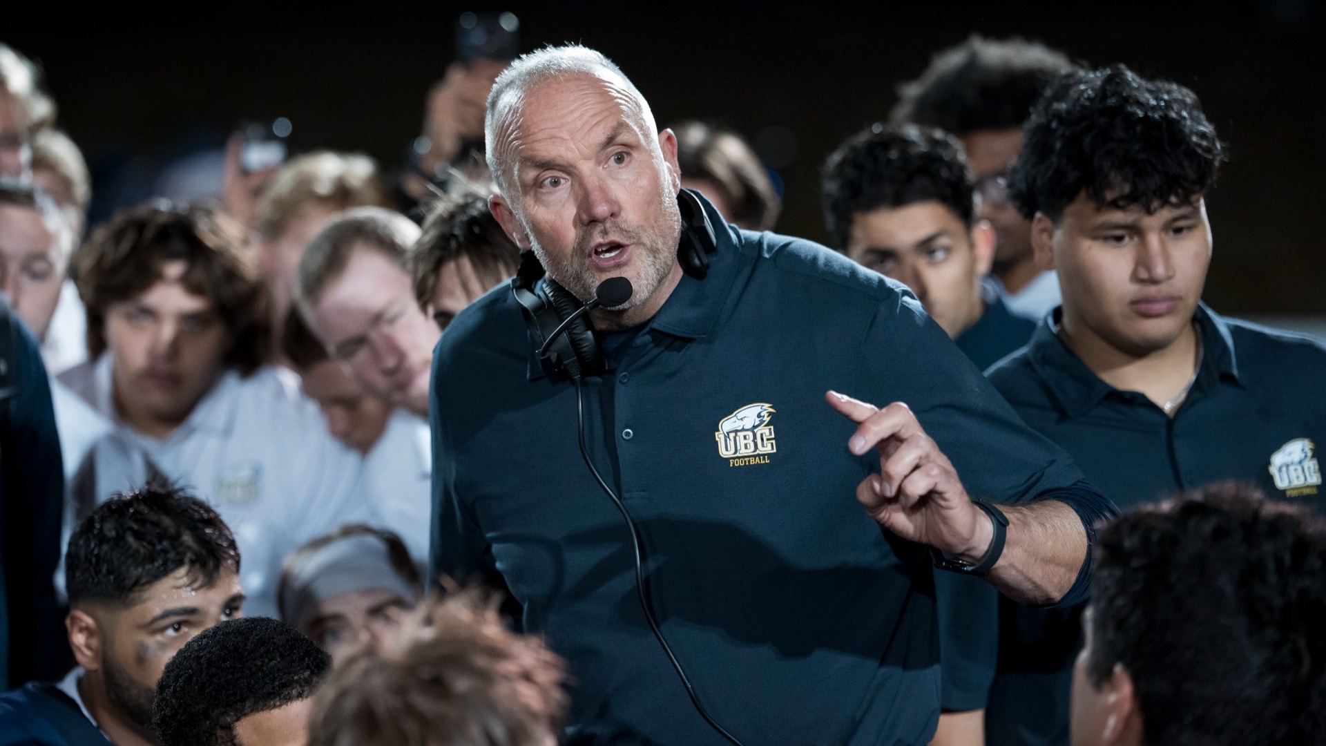 Blake Nill seen in semi-closeup, surrounded by players and staff, his headset down around his neck, speaking and looking intense
