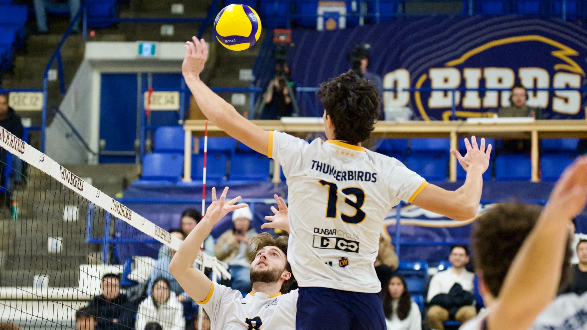 A UBC player is mid-air, winding up to swing at the ball over his head and above the net