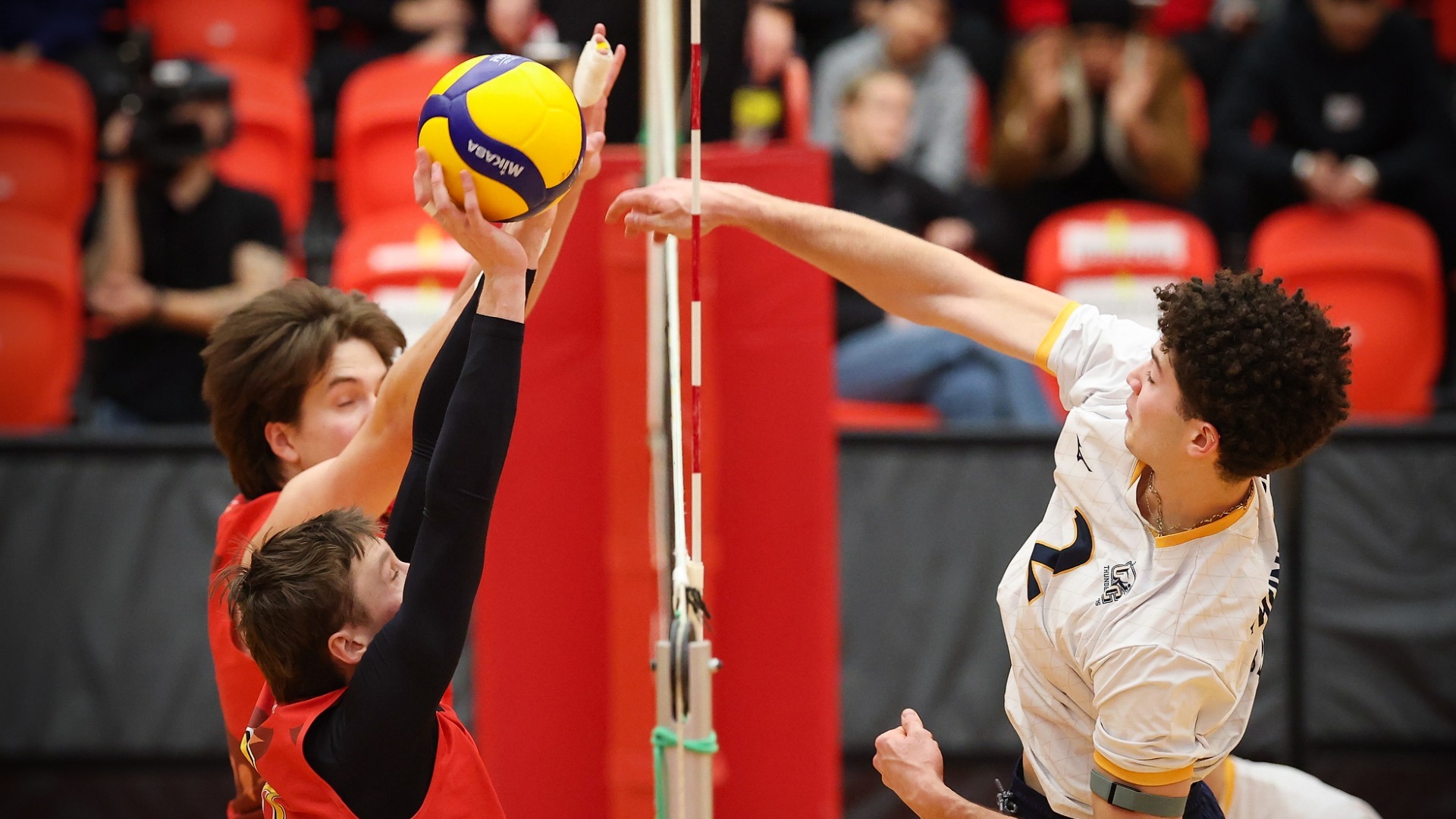 UBC Thunderbirds outside hitter Reeve Gingera following through on an attack, seen from top of the net. Two Calgary Dinos blockers attempt to turn him back, the ball is hitting the palm of one blocker, turning his wrist back.