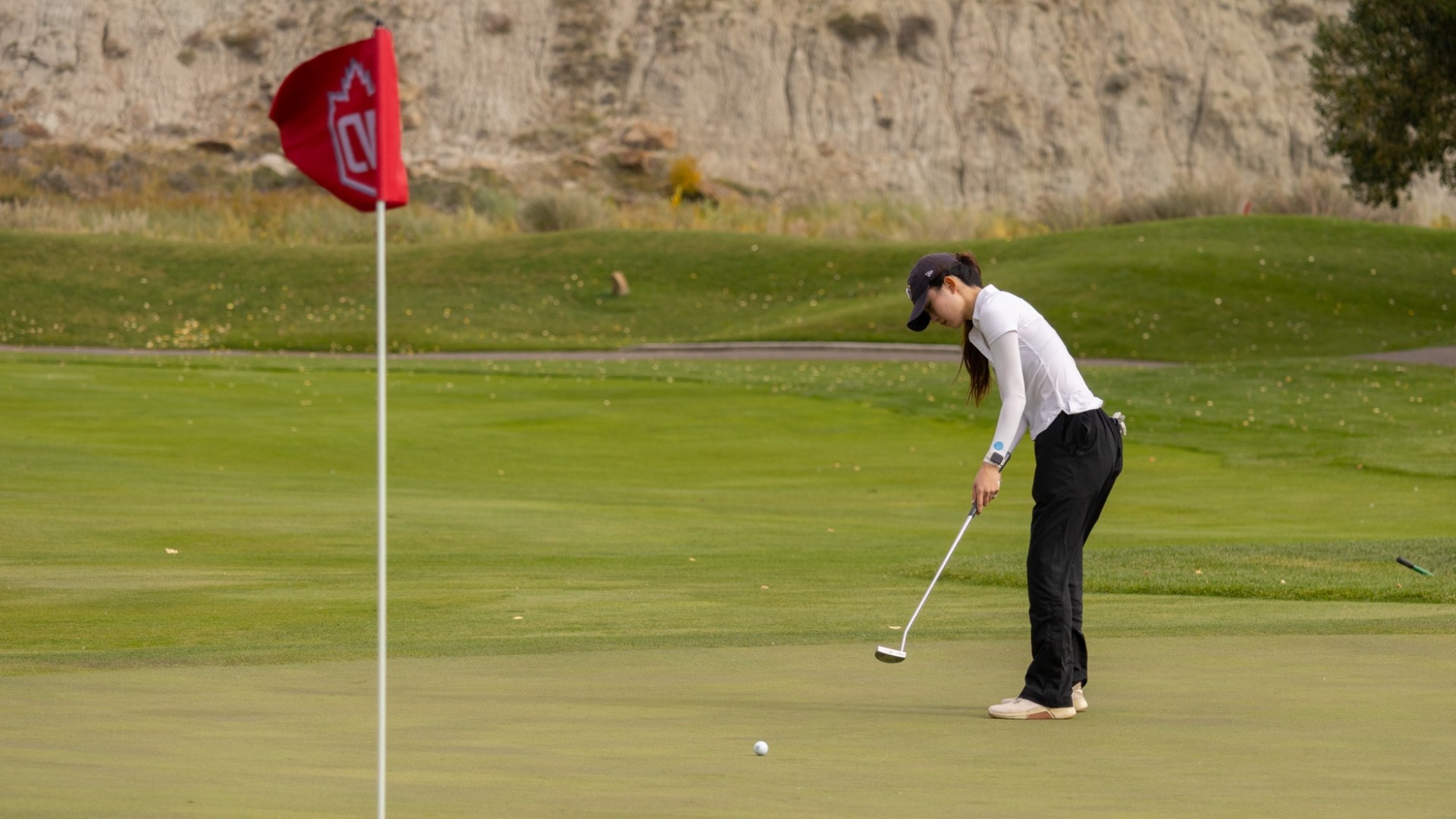 UBC Thunderbirds golfer Katherine Hao hits a long put toward the camera, with the red flag bearing a Canada West logo in foreground