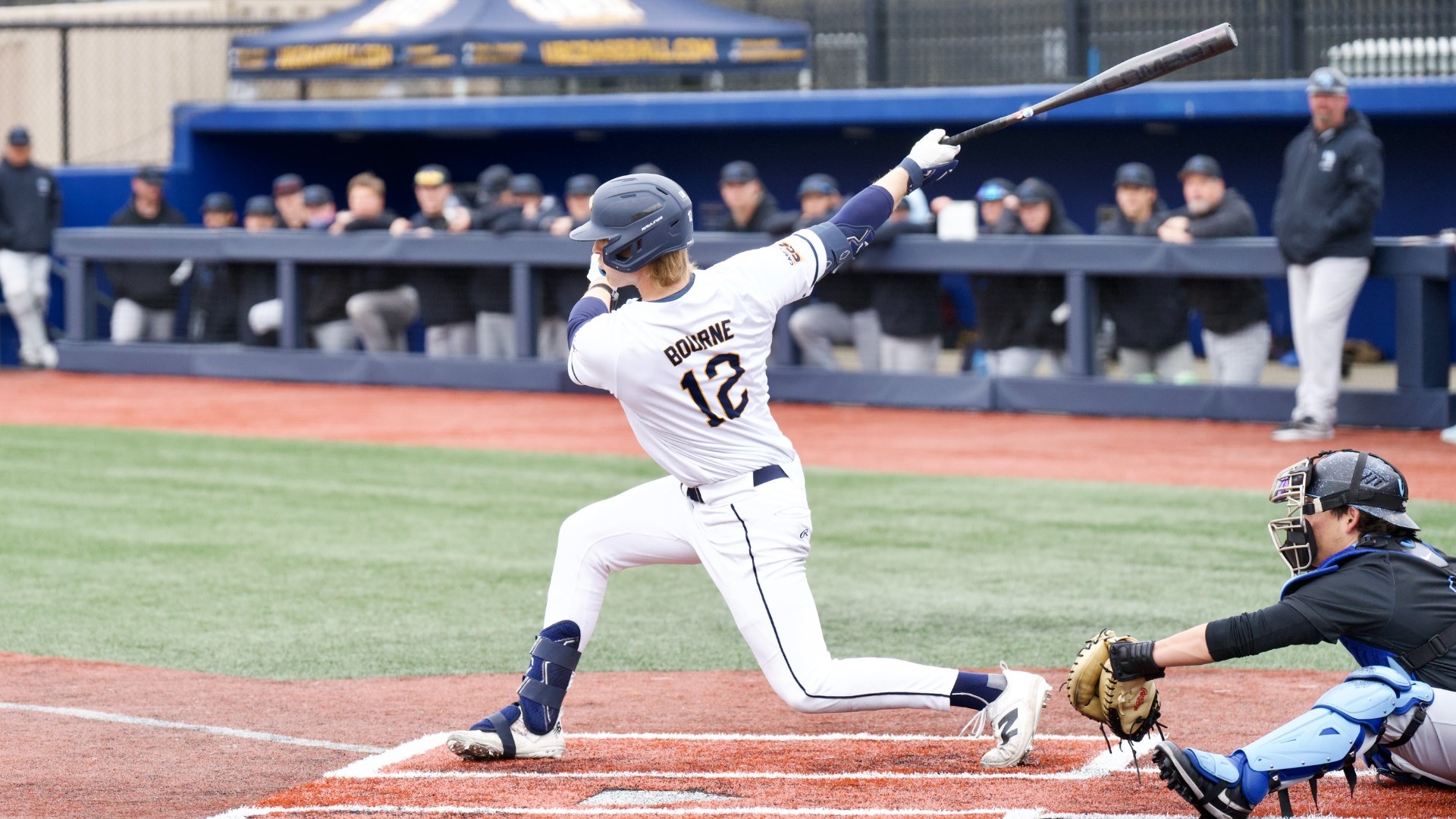 UBC hitter Kellen Bourne holding a long, one-handed follow through on his swing, watching the ball to his right from the left-handed batter's box