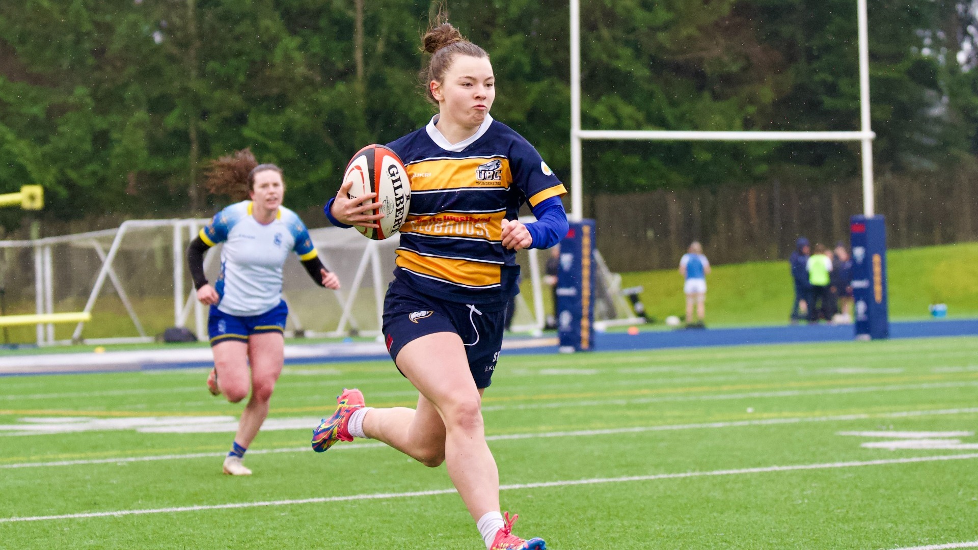 UBC Thunderbirds women's rugby 7s player Saylor Pickrell looking determined and perhaps a little sly as she sprints up the right side of the pitch with the ball. A Lethbridge Pronghorns player pursues, far behind.
