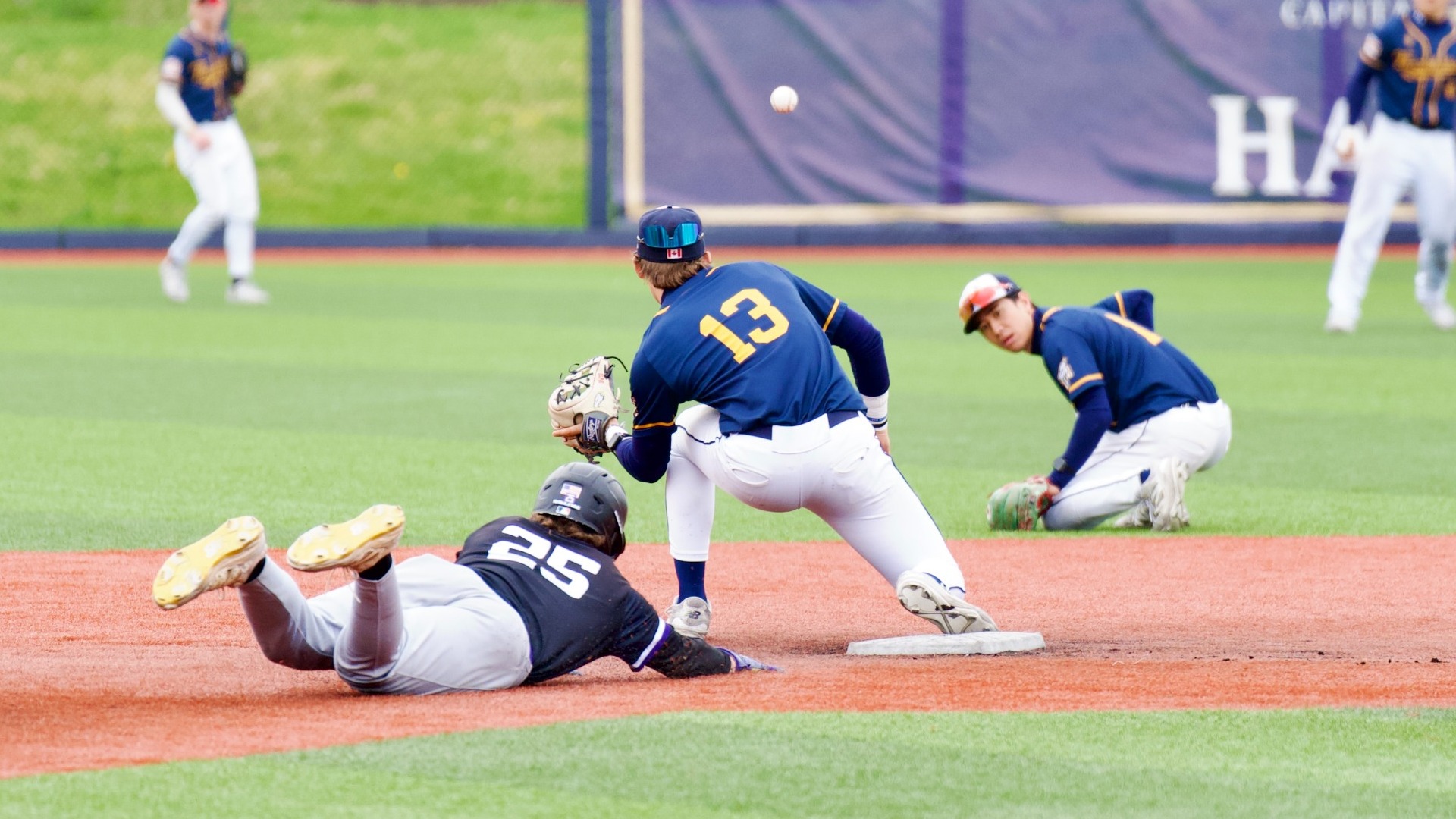 The UBC Thunderbirds making an out at second base. The shortstop reaching out for the ball, as the crouched second baseman crouches under a throwing from right field, watching the ball. A College of Idaho runner dives back to second base, but clearly won't beat the throw.