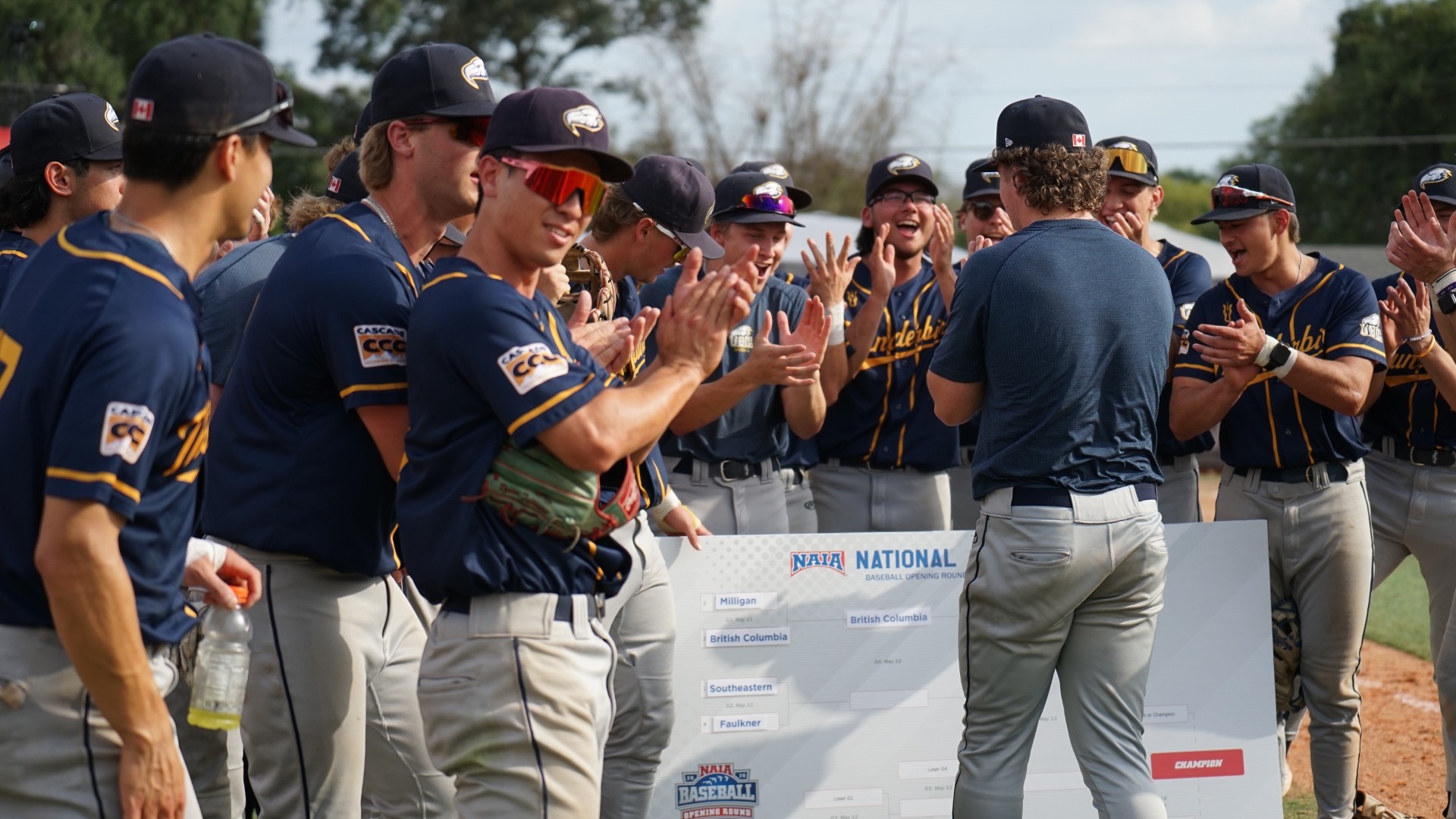 The entire UBC Thunderbirds baseball team clapping after starting pitcher Will Anderson moved the team forward on to the winner side of the bracket on an oversized board. Infielder Lou Fujiwara looks into the camera with a cool, half-smile as he claps. 
