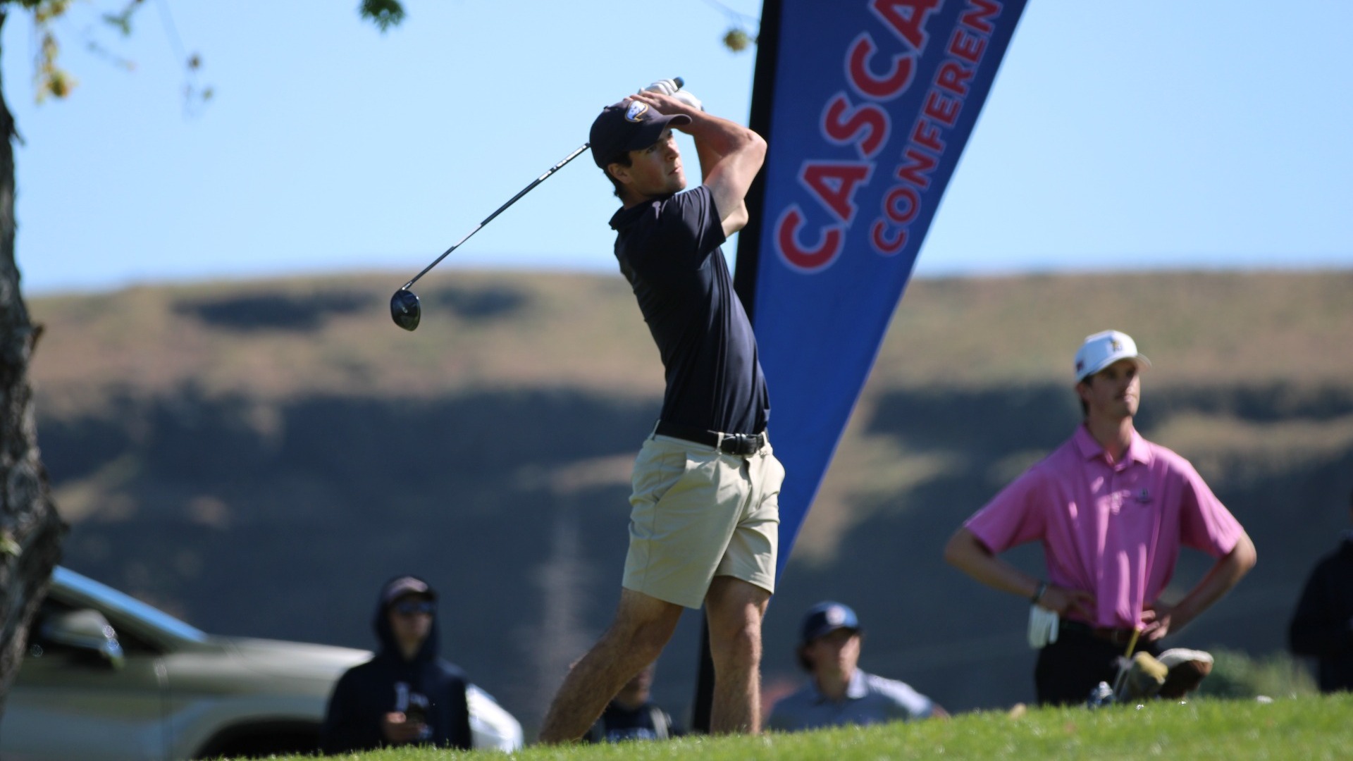 UBC Thunderbirds golfer Leighton Wright holding his hands high in the follow through of a drive, seen from a low angle with a Cascade Collegiate Conference flag billowing behind hium
