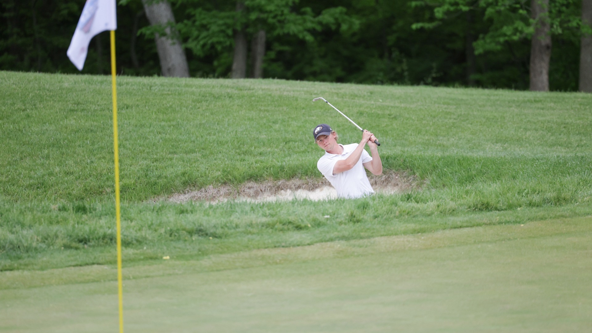 UBC Thunderbirds golfer Jake Houston hitting out of a bunker, toward the camera on the other side of the green. Only Houston's upper half is visible as he hold his follow through with his wedge above his head. The flagstick is out of focus in the foreground on the left side of the frame.