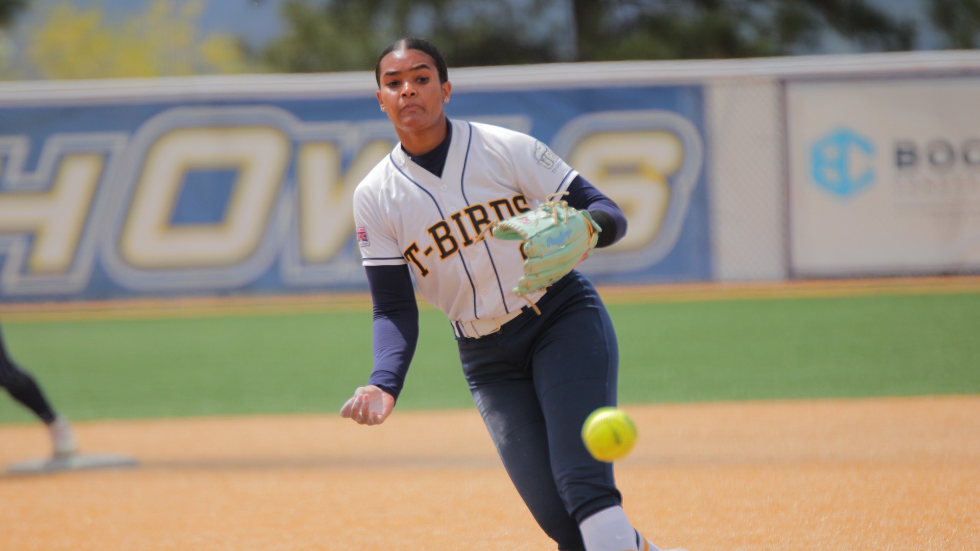 UBC Thunderbirds softball pitcher Carleen Murray delivering a pitch toward the camera, a serious look on her face, her hand down at her side just after releasing the pitch, the ball in the foreground and out of focus