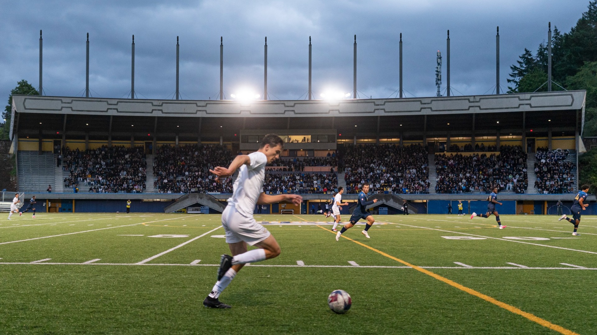 UBC Thunderbirds forward Max Comsia dribbling up the pitch, seen in the foreground of a fisheye wide angle shot capturing the full stands of Thunderbird Stadium behind him