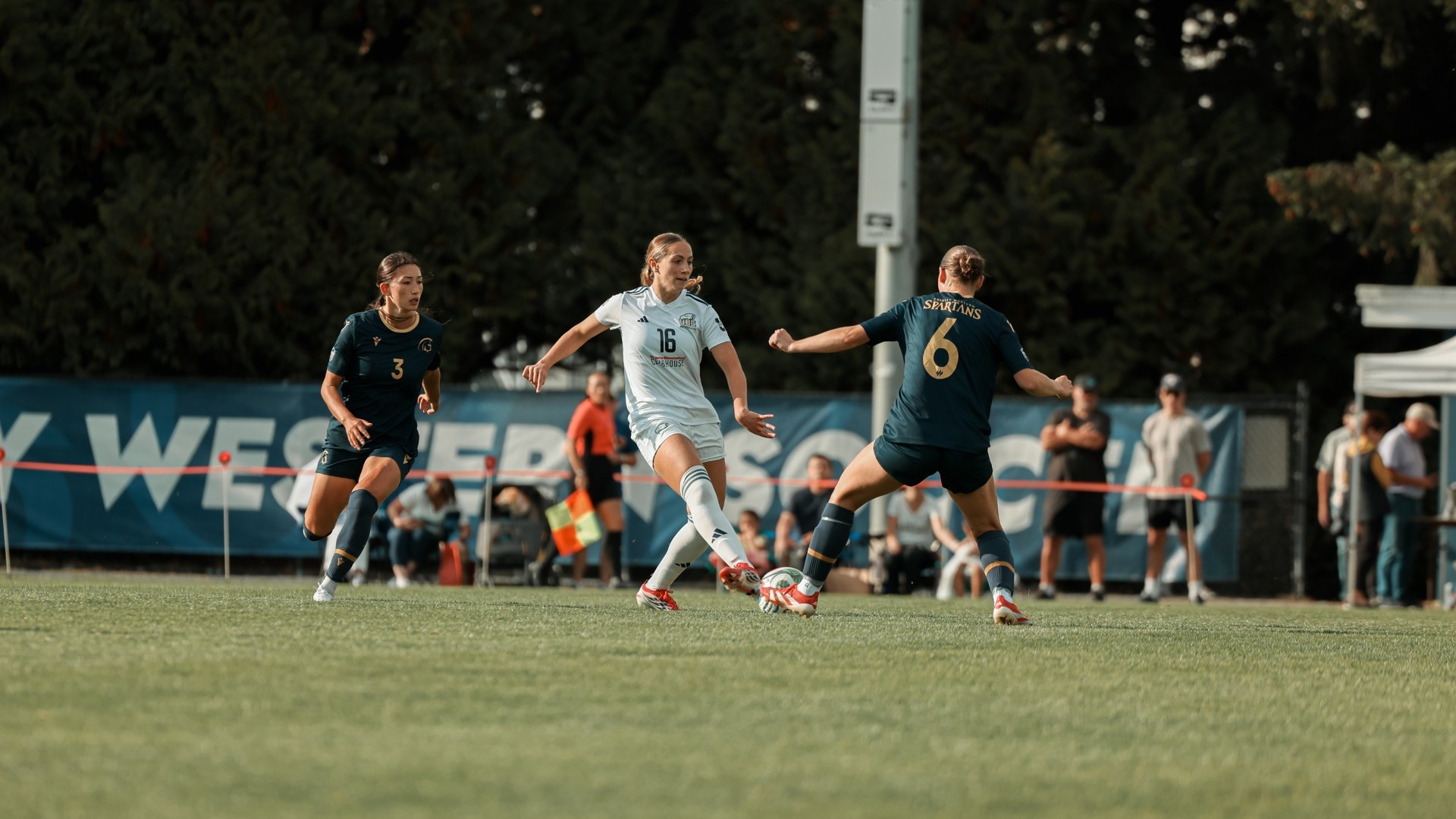UBC women's soccer player Felicia Hanisch makes a pass with her right foot as two Trinity Western Spartans players surround her, one lunging for the ball