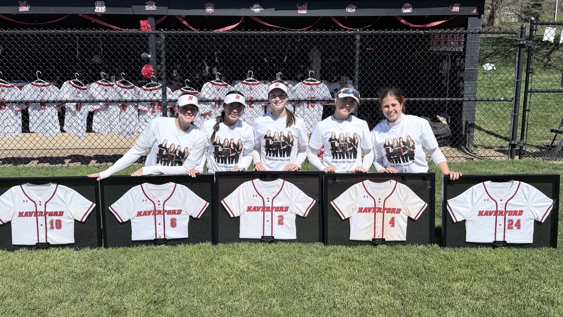 Five Haverford College softball seniors—Sarah Fan, Addie Sapirstein, Abby Litchfield, Jocelyn Leal, and Anna Stowe—stand shoulder to shoulder in front of the team dugout on Senior Day, smiling and wearing matching white T-shirts featuring a group photo. Each player rests a hand on a framed Haverford jersey displayed along the grass in front of them, with jersey numbers 10, 6, 2, 4, and 24 arranged left to right. Behind them, the dugout is decorated with red and black streamers and a row of hanging jerseys, creating a celebratory backdrop under bright sunlight.