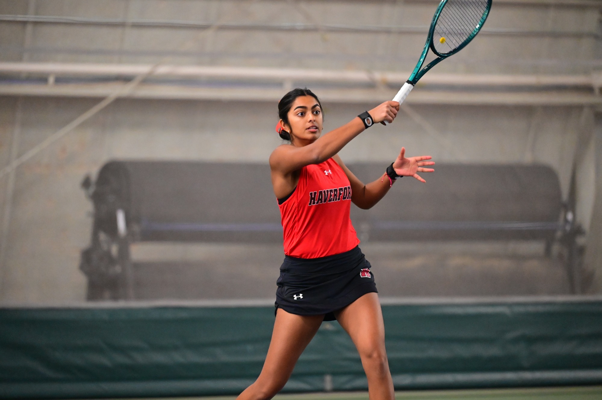 Action shot of Abhi in a red sleeveless jersey and black skirt preparing to strike a forehand, eyes locked on the ball. She holds her racket out in front with her right hand while her left hand is extended for balance. The indoor court background is softly blurred, with benches and a green backdrop visible behind her.