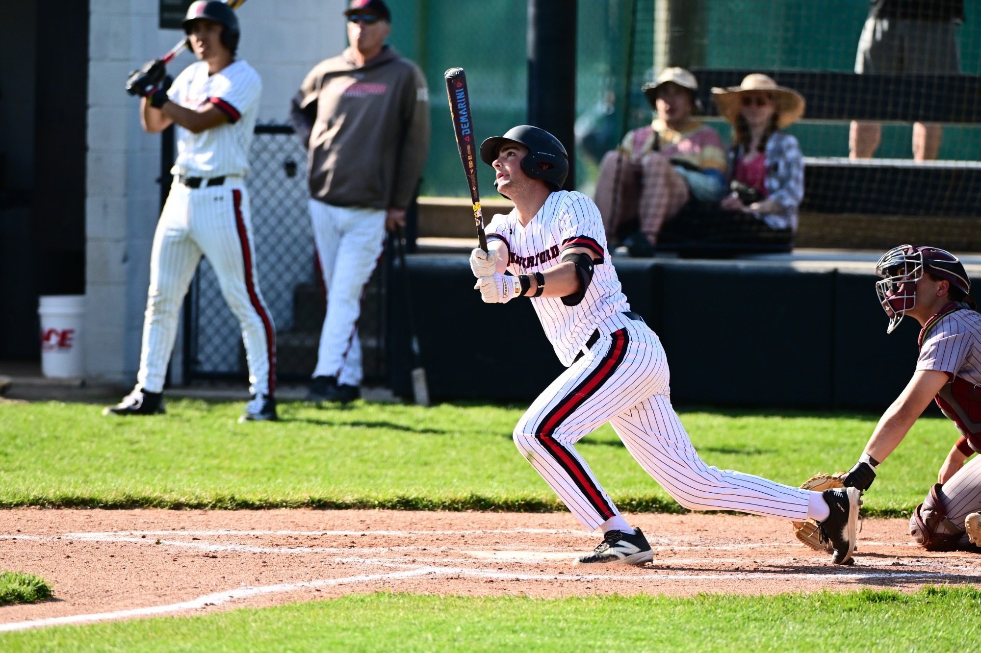 Right-handed batter Tommy Belman of Haverford watches the ball after making contact at the plate, holding his follow-through as he begins to step out of the batter’s box. A catcher in maroon gear remains low behind him, tracking the play, while a teammate on deck and a coach stand near the dugout in the background with a few spectators seated behind the fence. Bright sunlight casts sharp shadows across the infield dirt and grass.