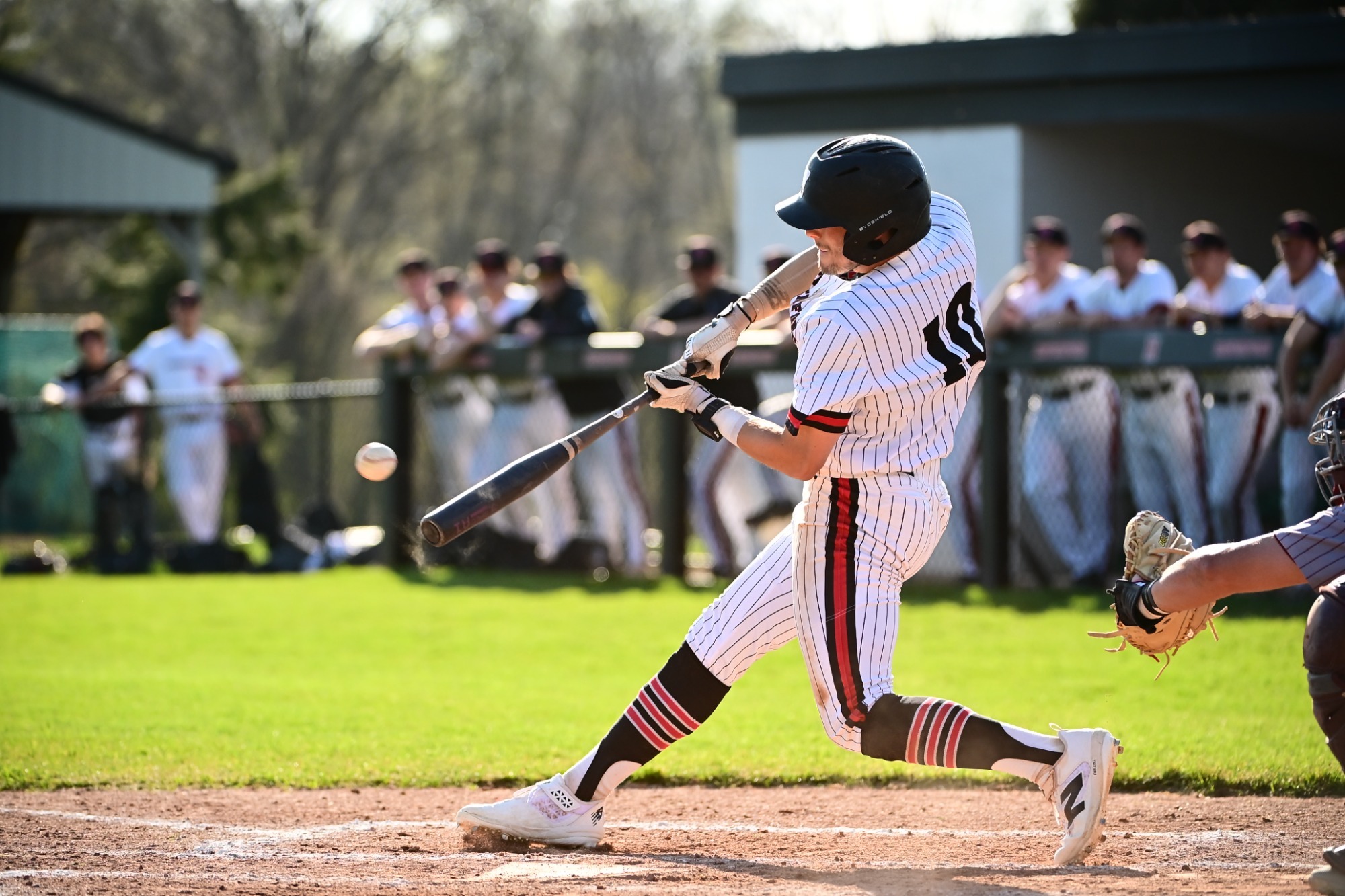  Haverford baseball player Luke Treese, wearing a white pinstriped uniform, connects for a powerful swing at home plate as the catcher looks on during a sunny home game.