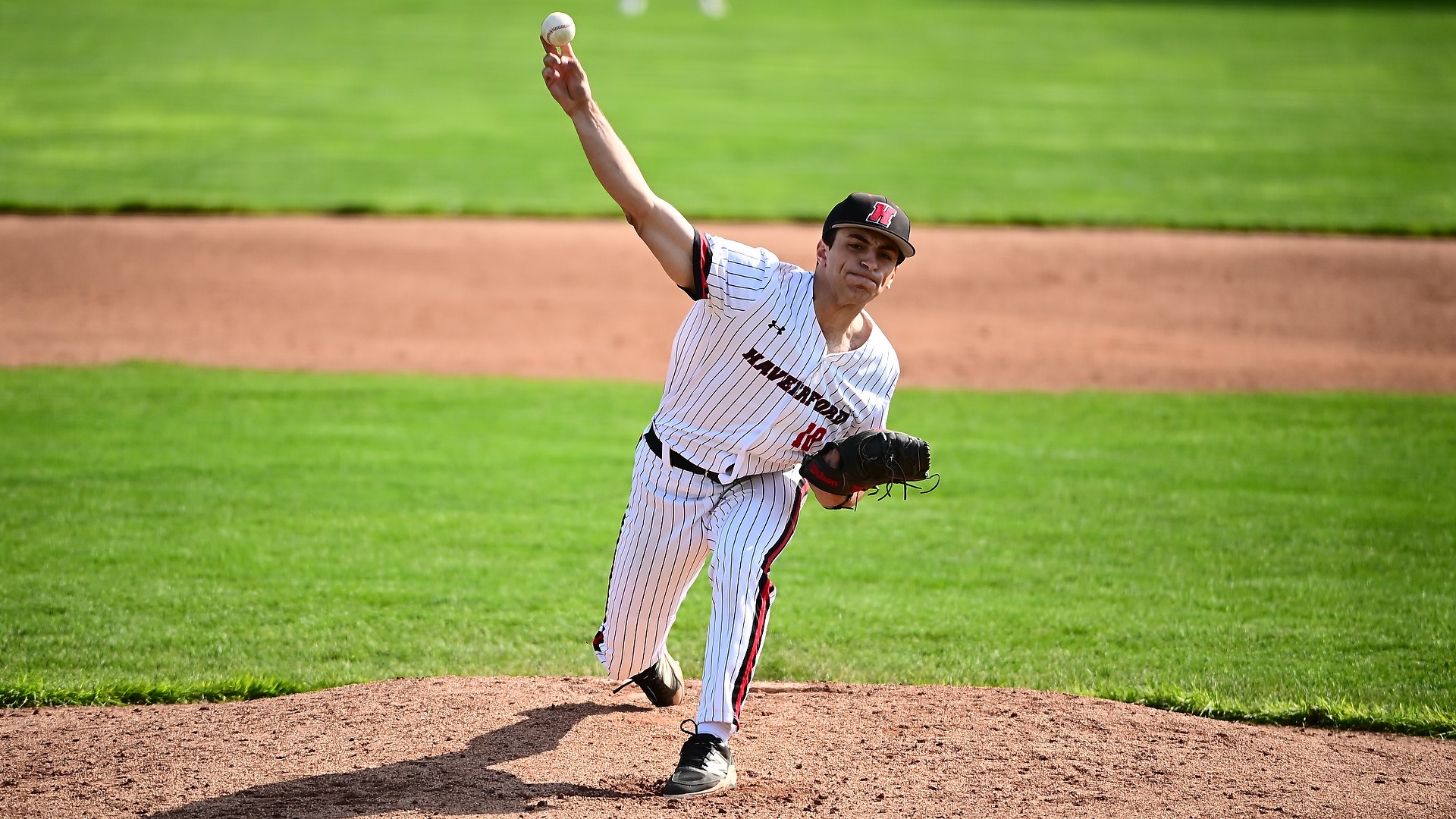 Right-handed pitcher Sammy Cohen delivers a pitch from the mound, fully extended in his throwing motion. He wears a white pinstriped Haverford uniform with a black cap featuring a red “H.” The baseball is just leaving his fingertips as his body leans forward, glove tucked near his waist. The dirt mound contrasts with the bright green outfield grass in the background under clear daylight.