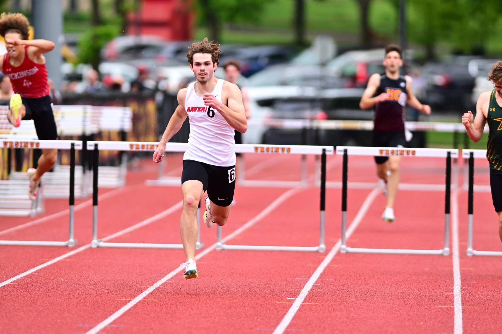 Haverford College track athlete Nico Greene sprinting toward the camera on a red outdoor track. He is wearing a white Haverford singlet with the number 6 and black shorts. In the background, other runners are visible, including one leaping over a hurdle on a parallel lane during a track meet.